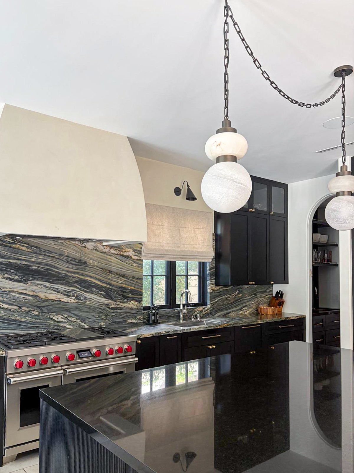 Moody black kitchen with quartzite countertop and backsplash, granite countertop, plaster hood, polished nickel and brass hardware, stainless steel range, bronze sconces, and alabaster pendant lights.