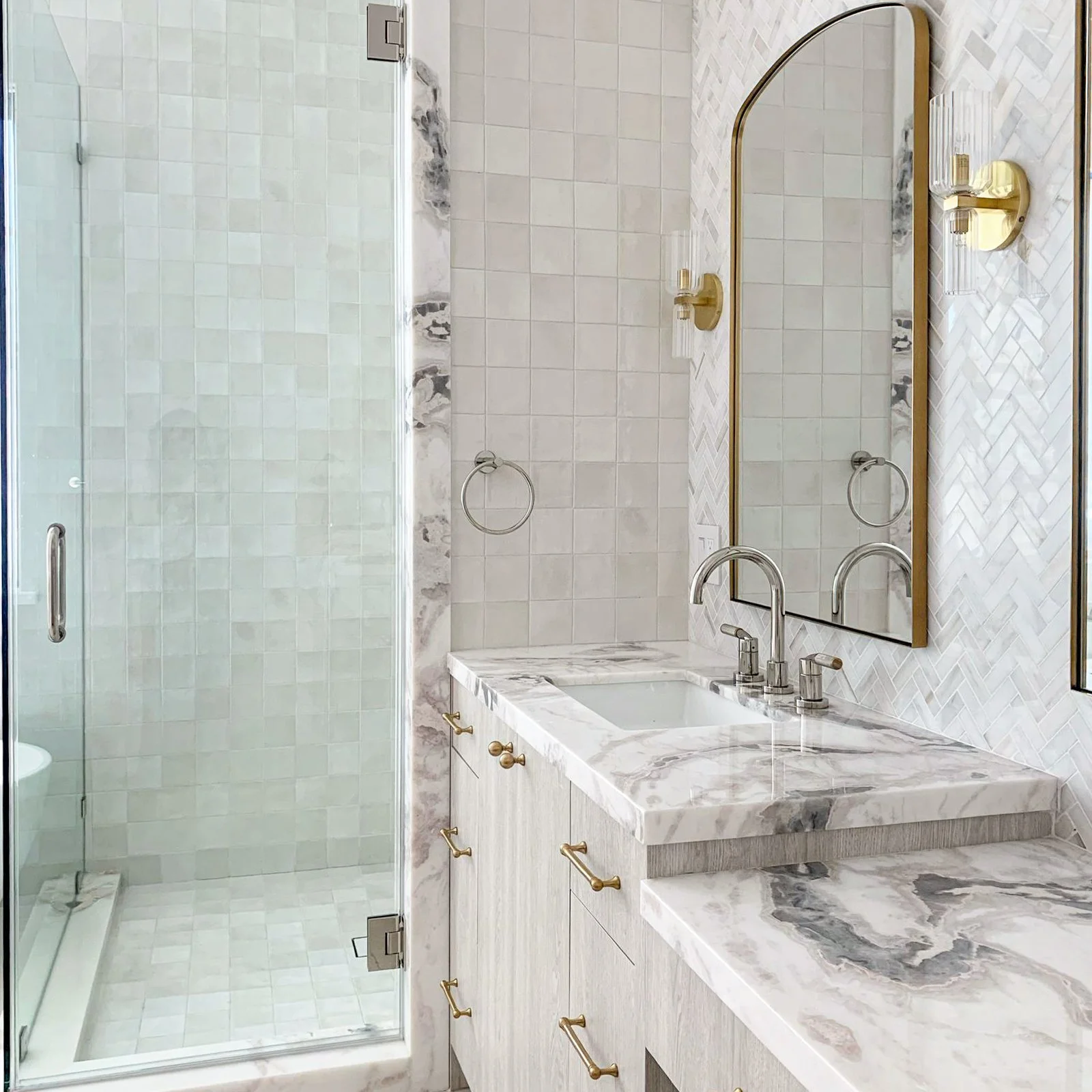 Primary bathroom in cream and gray tones, with antique brass and polished nickel finishes, arched mirrors, tiled shower and vanity wall, marble countertop and accents, and freestanding soaking tub.