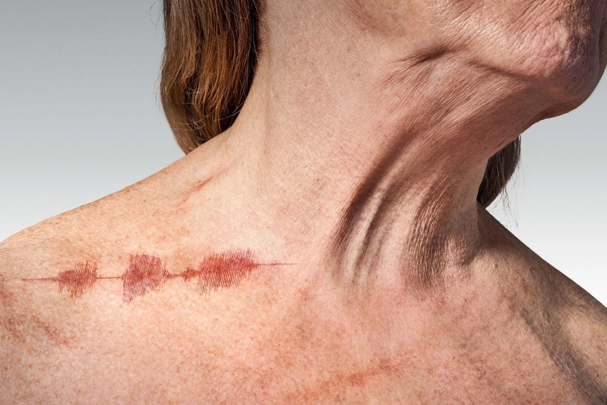 Close-up of a woman's neck, shoulder, and lower face showing visible wrinkles, a scar, and a tattoo of a sound wave on her shoulder.