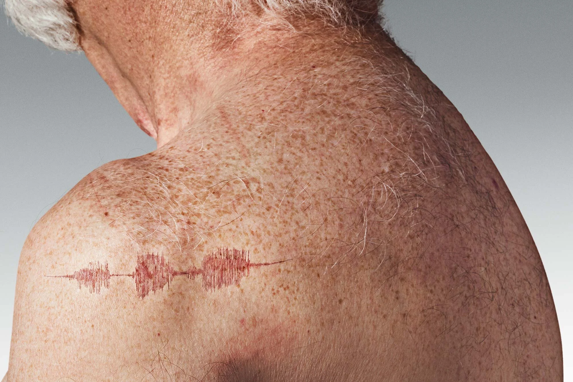 Close-up of an older man's shoulder and neck area with freckles, gray hair, and a red tattoo of a sound wave pattern.
