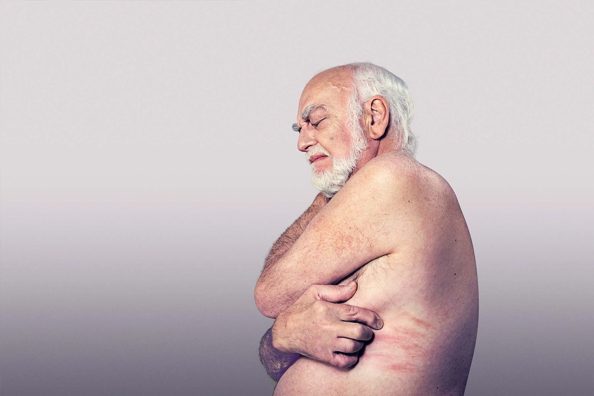 An elderly man with a white beard and hair, appears to be topless and is standing with his eyes closed and a pained expression, hugging himself against a plain light-colored background.