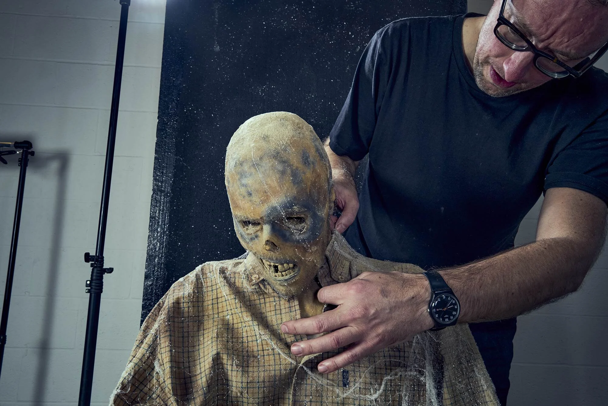 A person wearing glasses and a black t-shirt holds a zombie-like puppet with a decayed face, exposed teeth, and hollow eyes, in front of a dark backdrop in a studio setting.