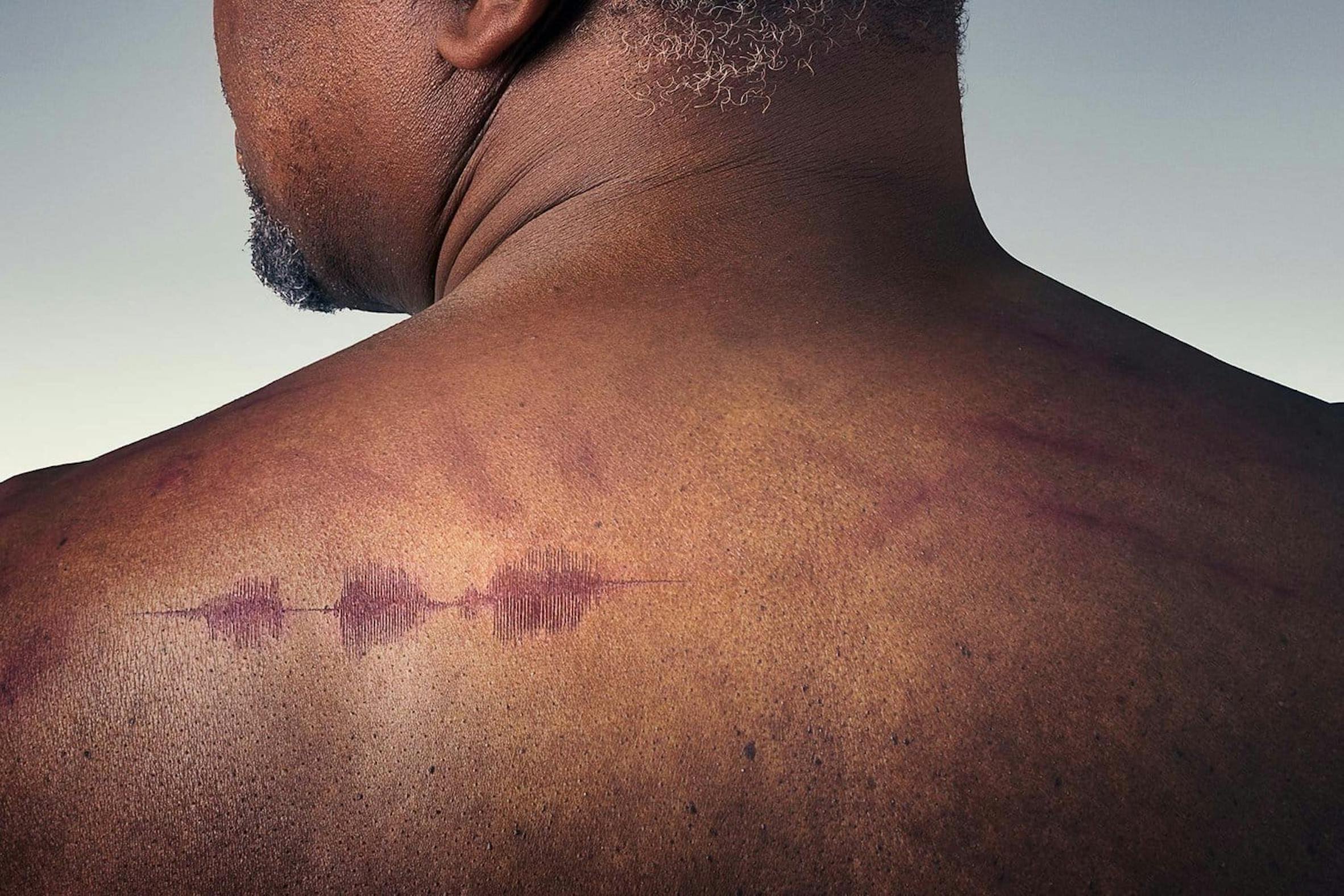 Close-up of a man's shoulder and neck, with a visible sound wave tattoo on the shoulder.