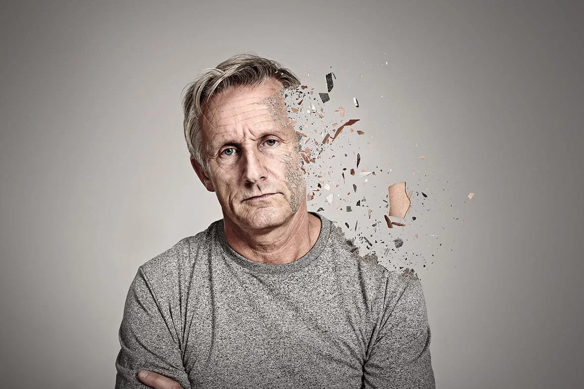 A middle-aged man with gray hair in a gray shirt appears to have part of his face and head disintegrating into dust and debris, giving the impression of a digital or artistic explosion.