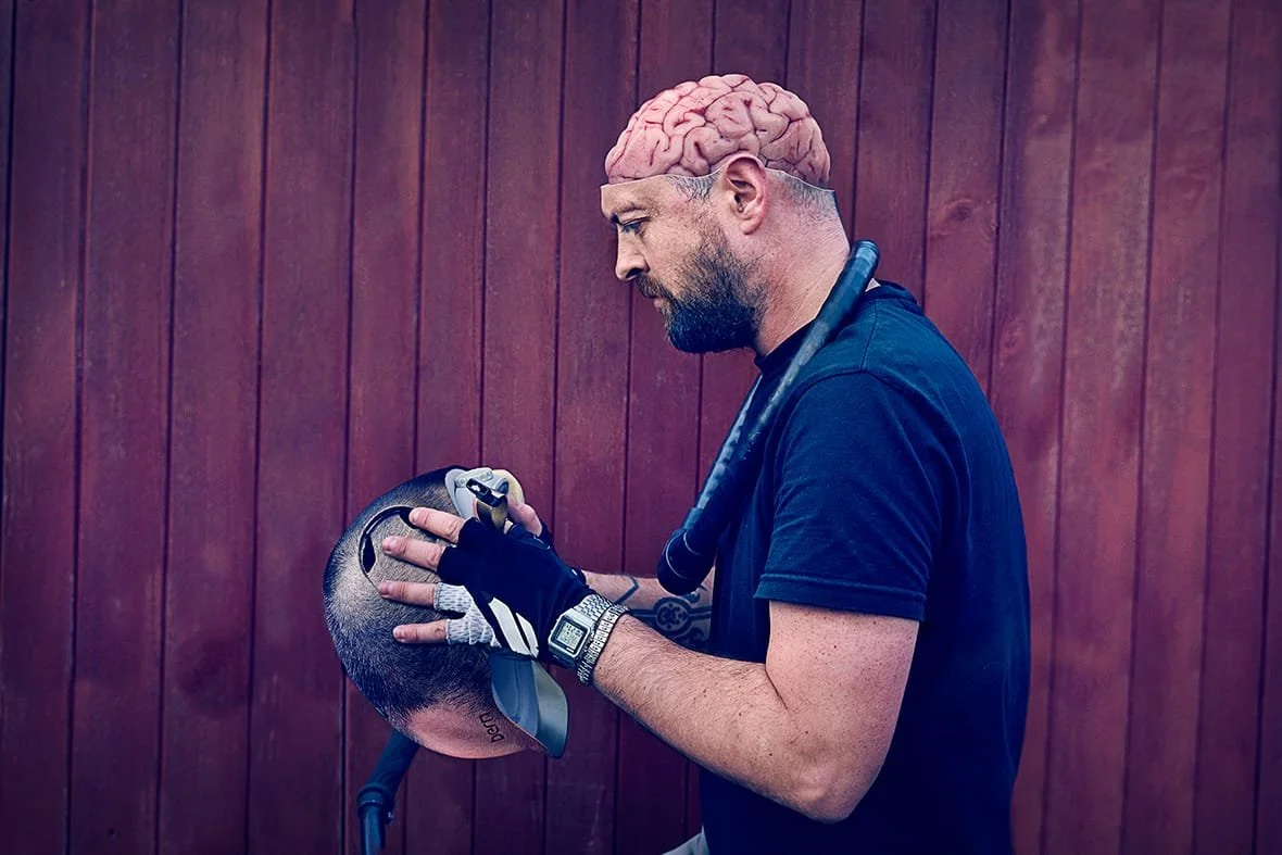 A man with a shaved head and beard, wearing a black shirt,, holding a bike helmet, with a pink brain graphic on his head, standing against a wooden wall.