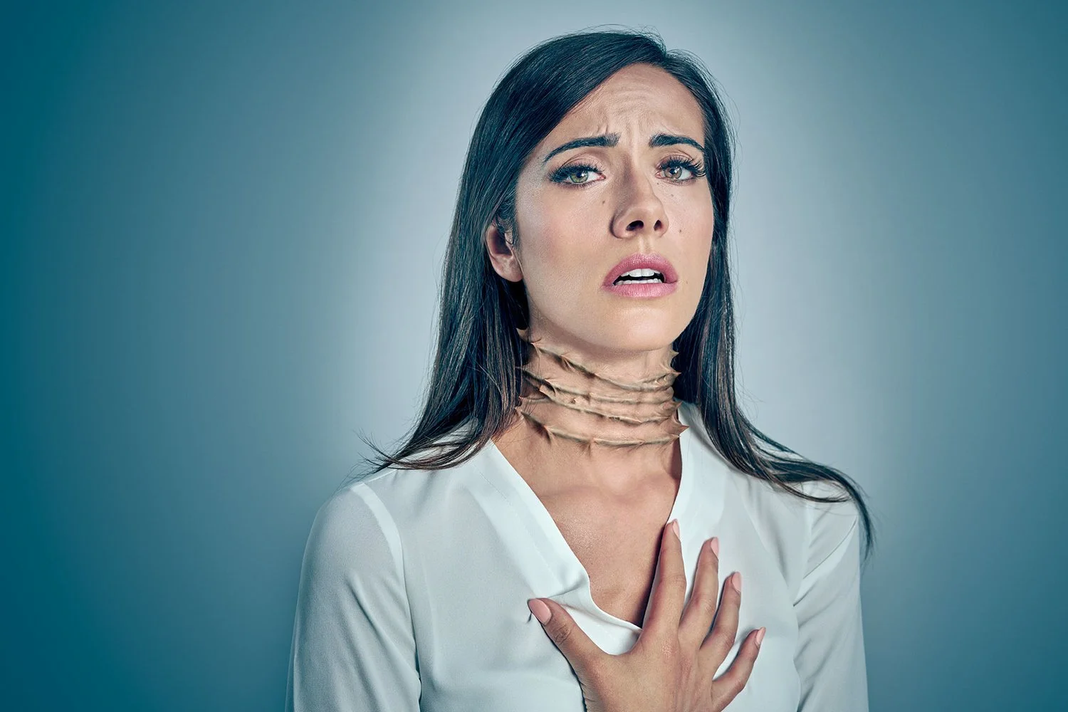 A woman with dark brown hair and light makeup appears distressed, holding her chest with her left hand. She has a large, flesh-colored, multi-layered choker around her neck, and she is wearing a white top. The background is plain and gray.