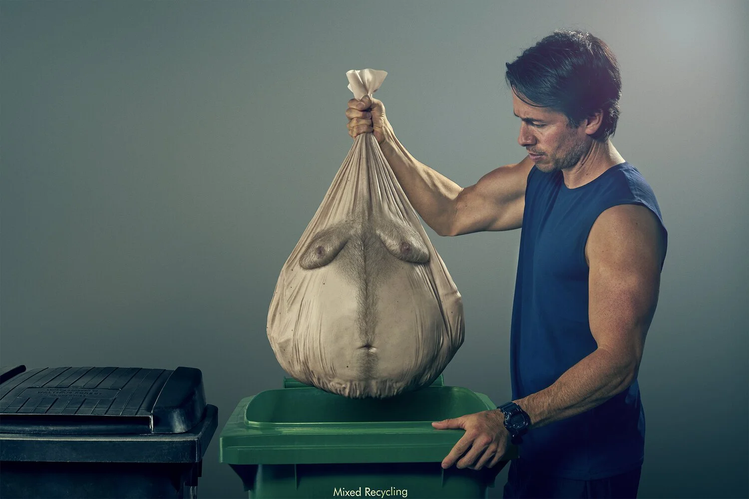 Man holding a large bag of mixed recyclable waste with a waistband of a woman's torso printed on the bag, placed in a recycling bin.