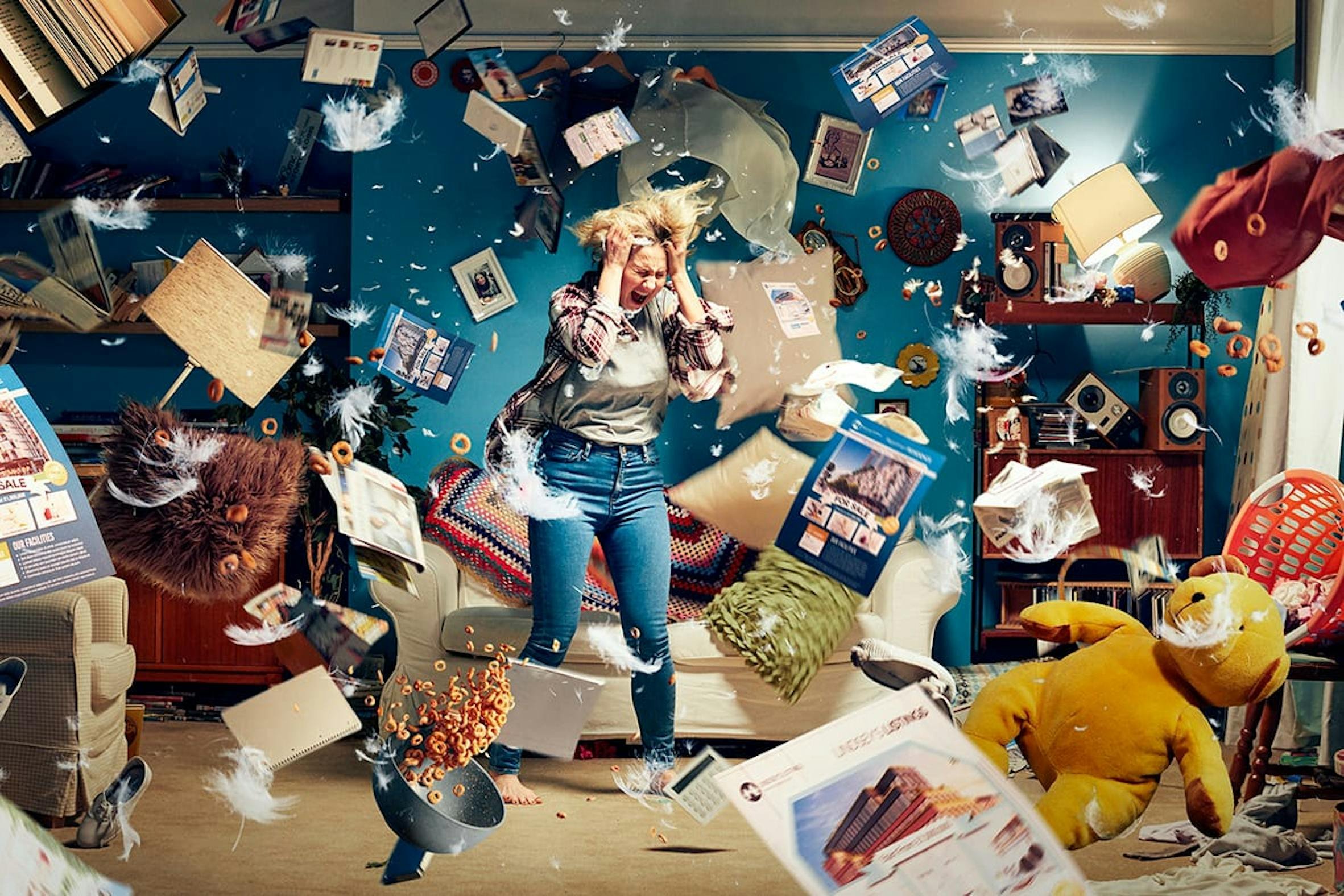 A woman standing in a living room during a chaotic mess, surrounded by flying papers, feathers, and cereal, with a distressed expression, in front of a sofa and cluttered shelves.