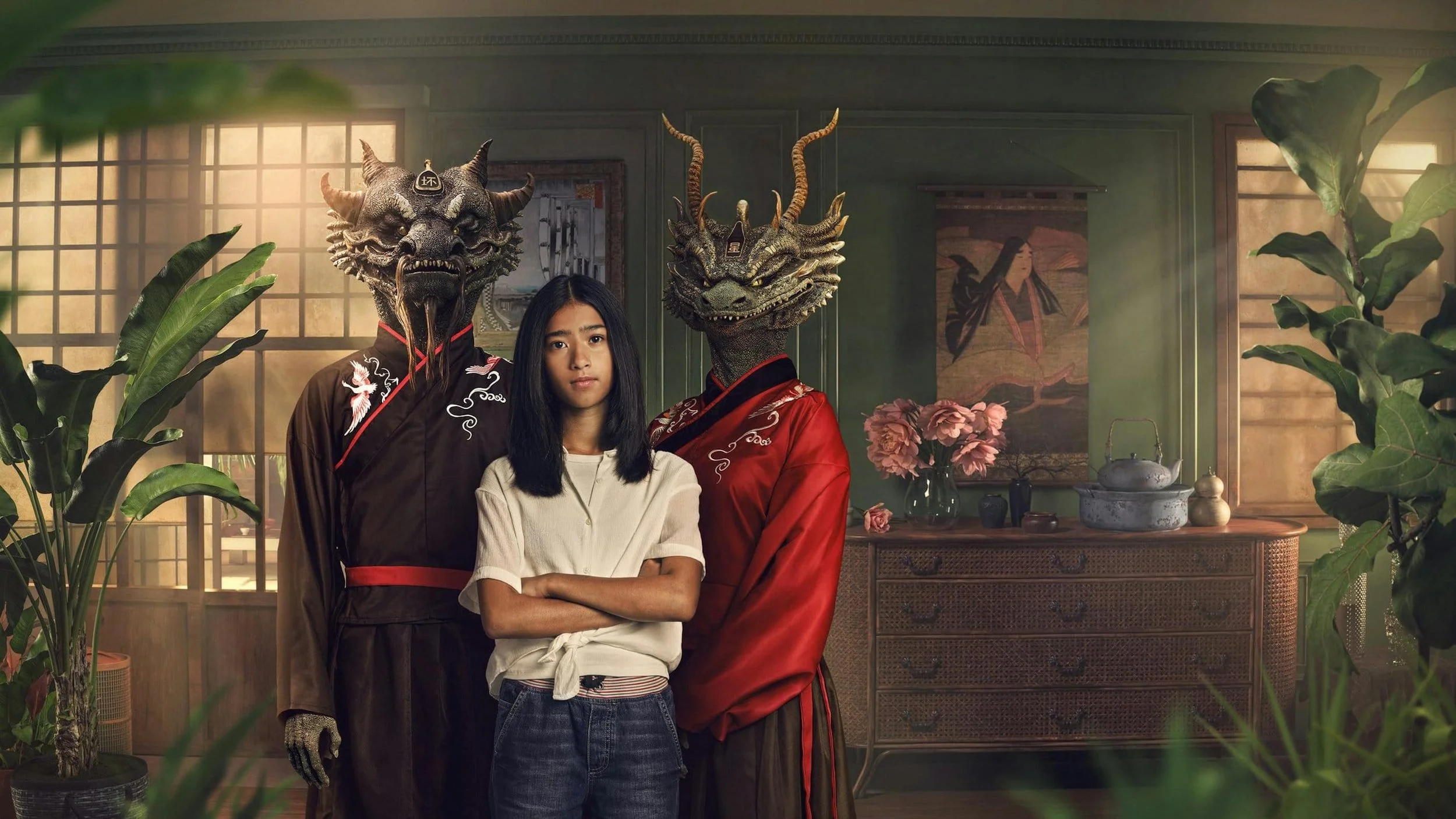 A young girl with crossed arms standing between two individuals wearing dragon masks and traditional Asian clothing, inside a room with plants and Asian decor.