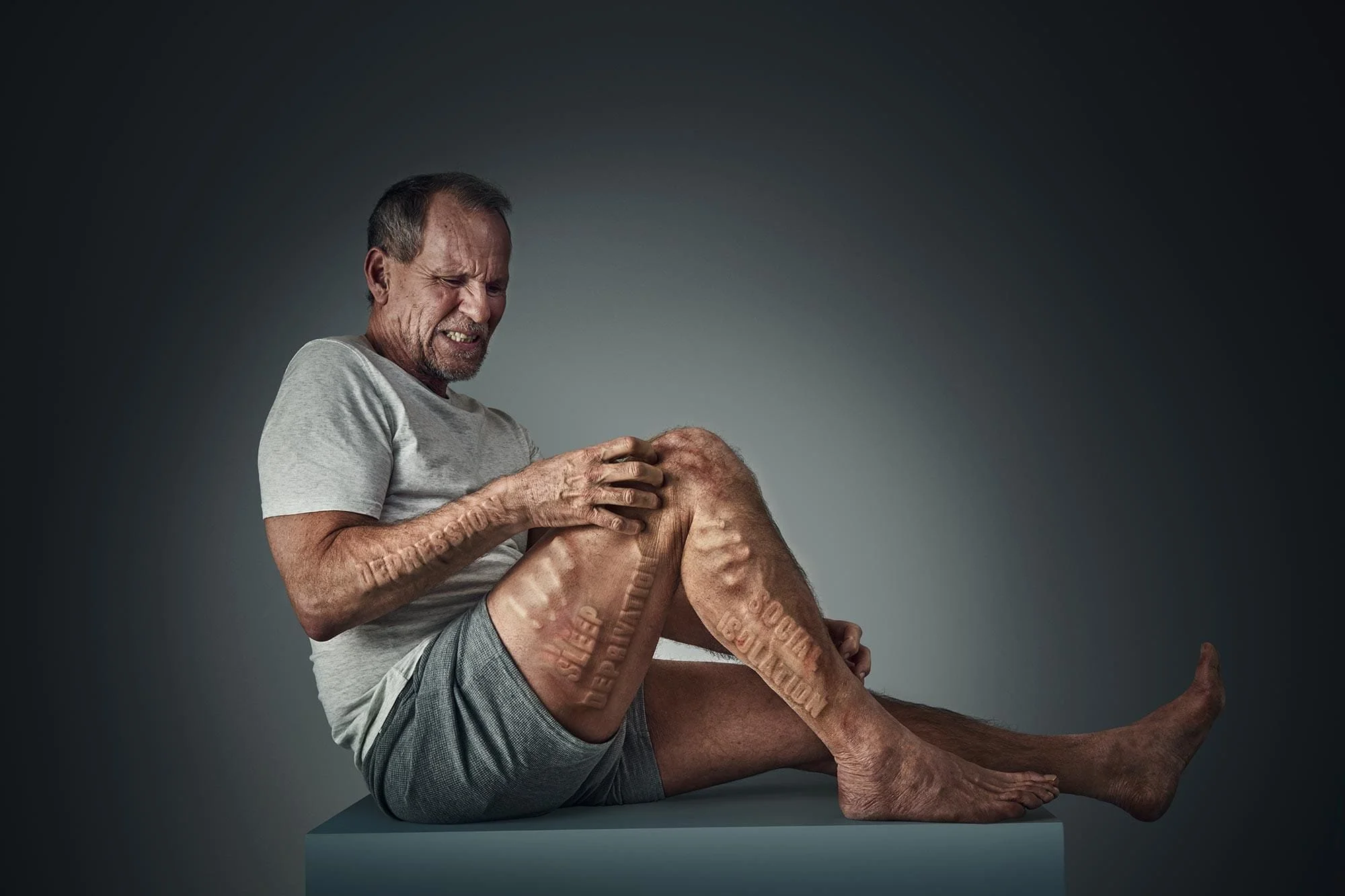 A man with a pained expression sitting on a platform, holding his knee, which has words like 'sleep,' 'stress,' 'deprivation,' and 'anxiety' engraved on it.