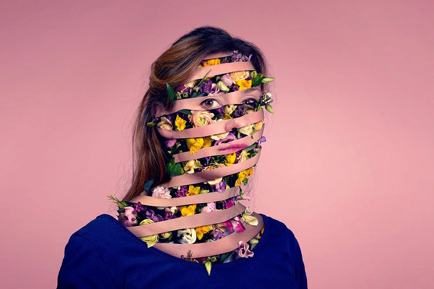 A person with a face made of various colorful flowers and ribbons wrapped around their head and face, against a pink background.
