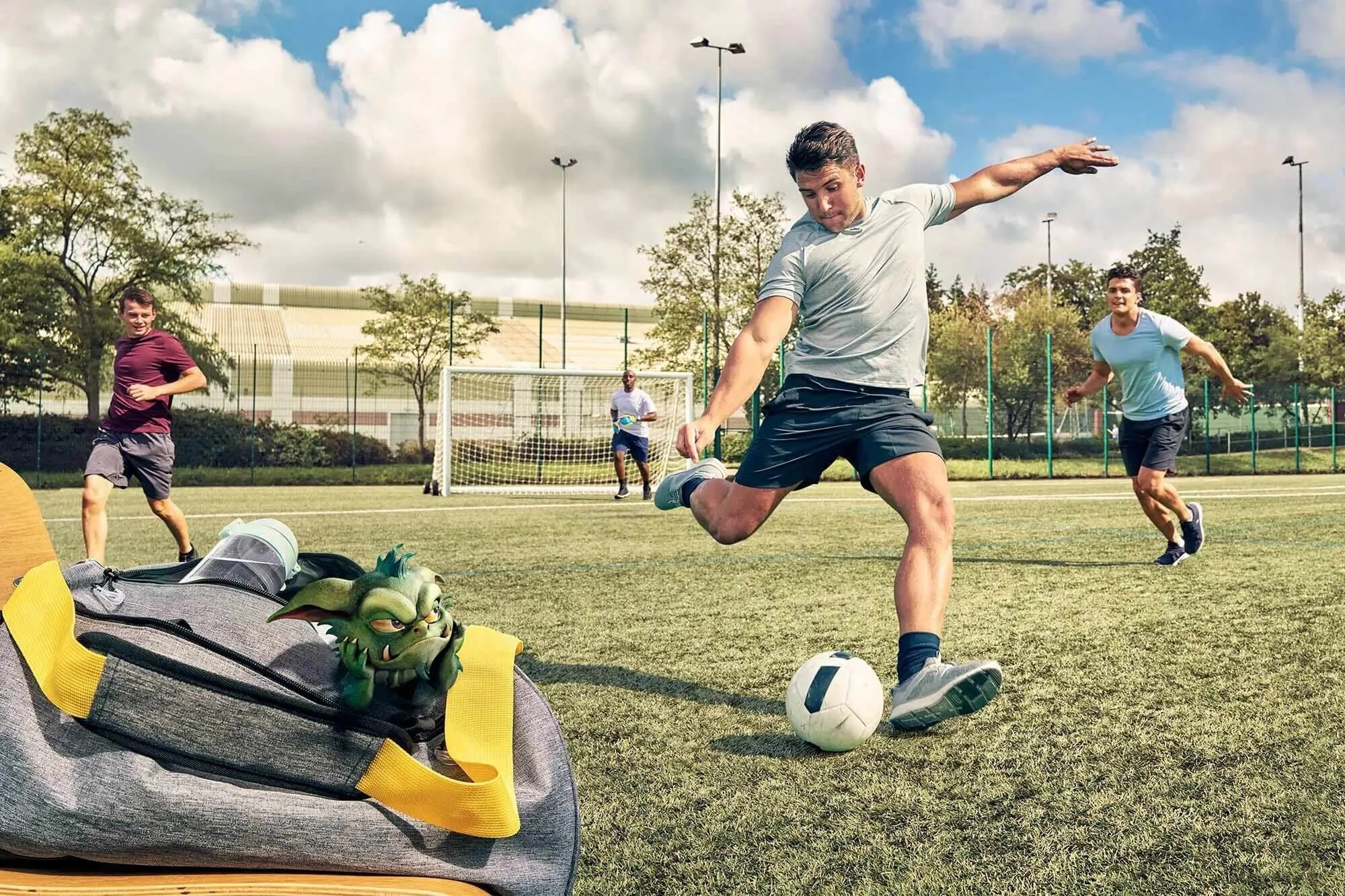 A group of young men playing soccer on a field, with one man kicking the ball while others run behind him. There is a backpack on the ground with a green dragon toy on top of it, and a soccer goal in the background.
