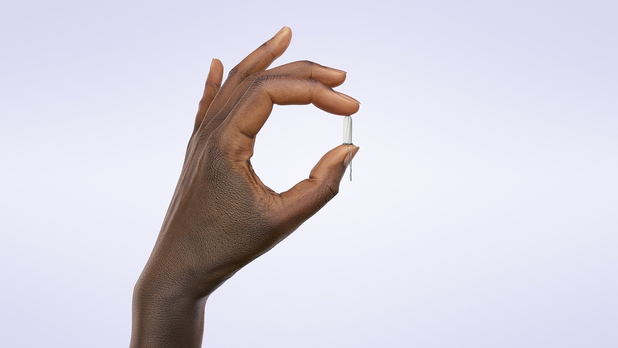 A hand with dark skin holding a white capsule pill between thumb and index finger against a plain light background.