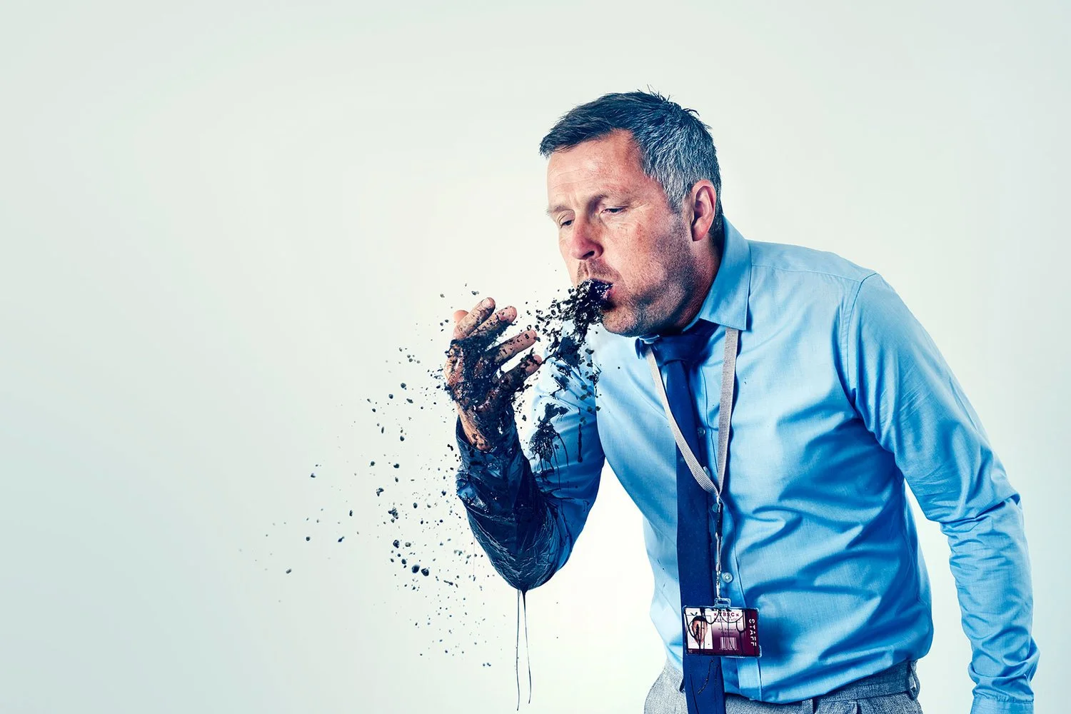 A man in business attire with a badge on his neck is coughing into his hand, which is covered in black ink or paint, causing a spray of ink or paint to disperse as he coughs.