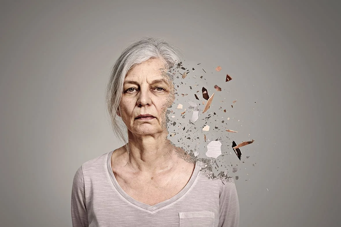 A woman with gray hair and a somber expression has part of her face and neck dissolving into scattered debris and fragments, creating a digital or artistic effect.