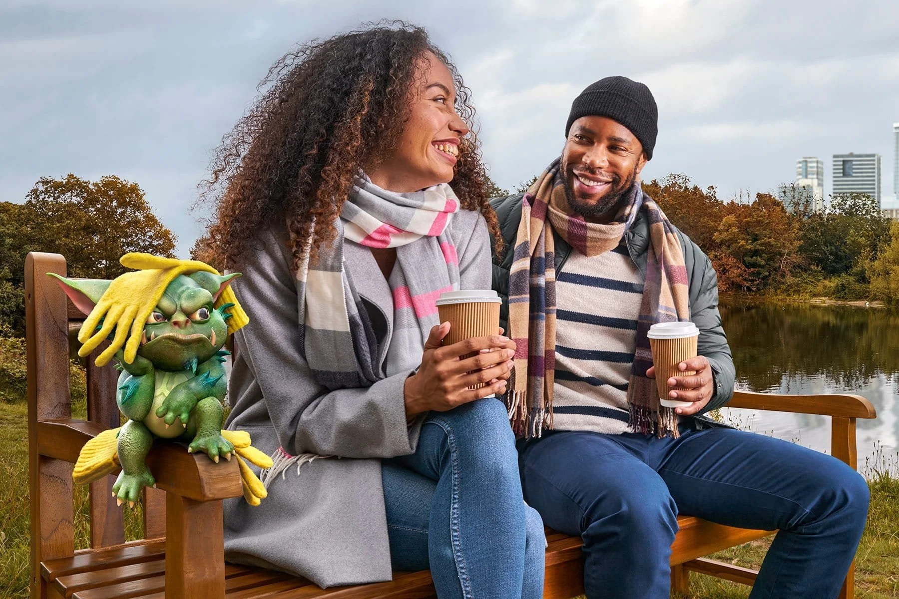 A woman and a man sitting on a park bench, smiling and holding coffee cups, with animated plush toy of a green creature with yellow hair sitting beside them. They are outdoors near a lake with autumn trees and city buildings in the background.