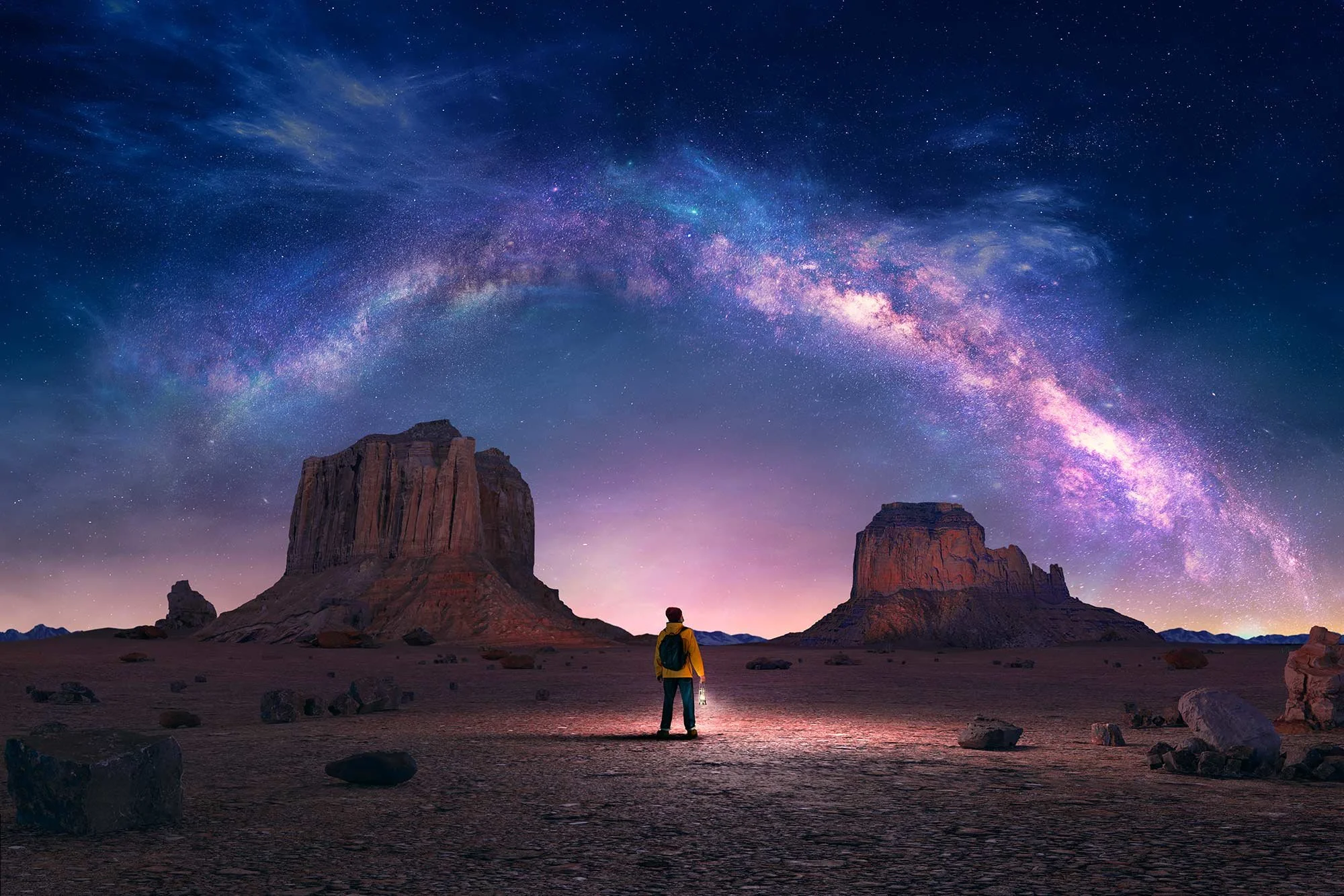A person with a backpack holding a lantern, standing in a desert at sunset, with two large rock formations and the Milky Way galaxy arcing across the sky.