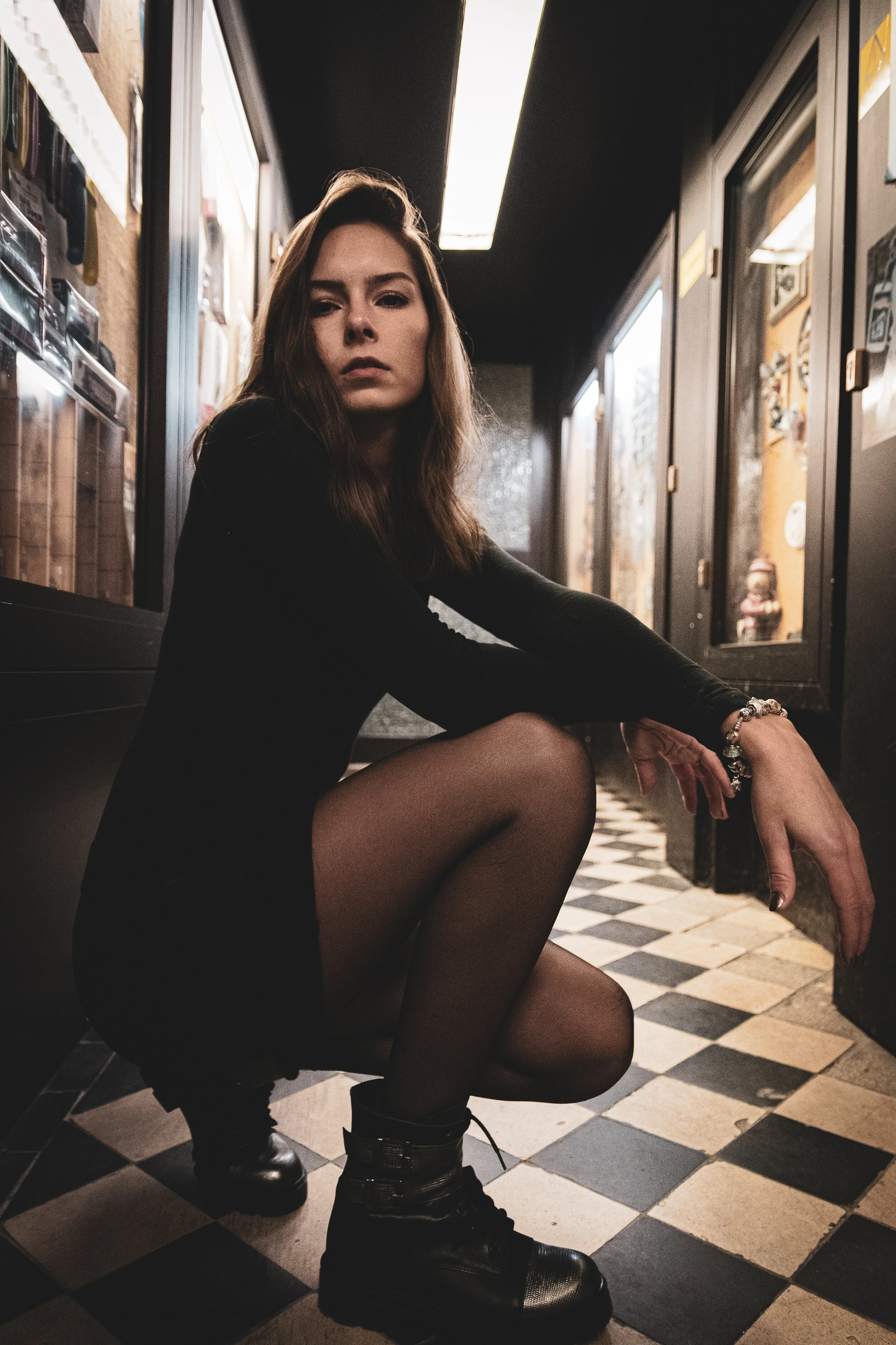 Eine junge Frau mit braunen Haaren sitzt in einem Flur mit schwarz-weißer Fliesenboden, gekleidet in einen schwarzen Pullover, schwarze Kleidung, Strümpfen und Stiefeln, die auf dem Boden stehen, schaut ernst in die Kamera.