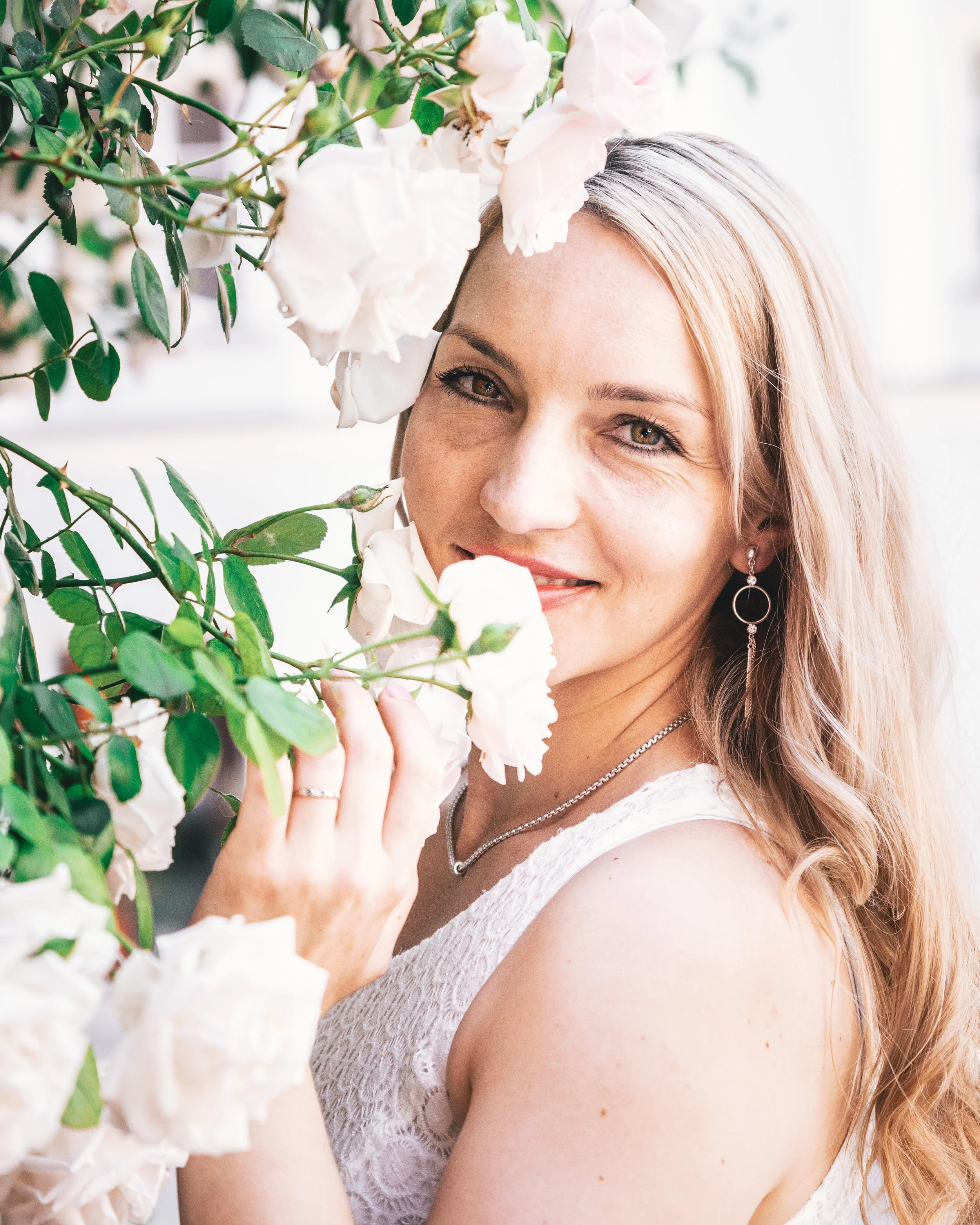 Eine lachende Frau mit langen blonden Haaren, die zwischen weißen Rosenblüten steht, trägt ein weißes Kleid und silbernen Schmuck.