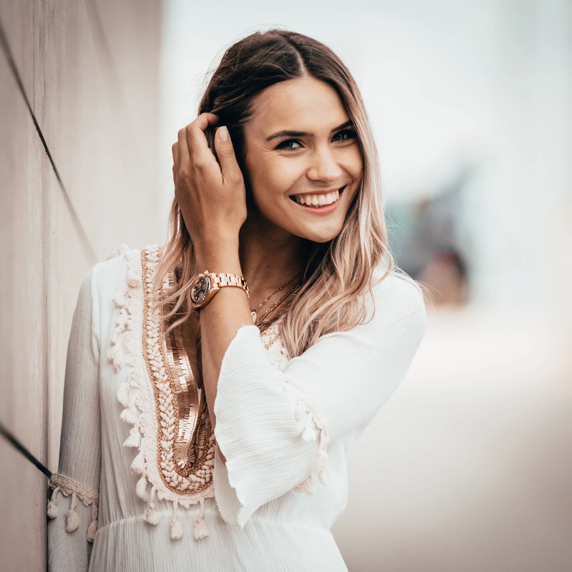 Eine lachende junge Frau mit langem, gewellten Haar, trägt eine weiße Bluse, einen goldenen Schmuck und eine Armbanduhr.