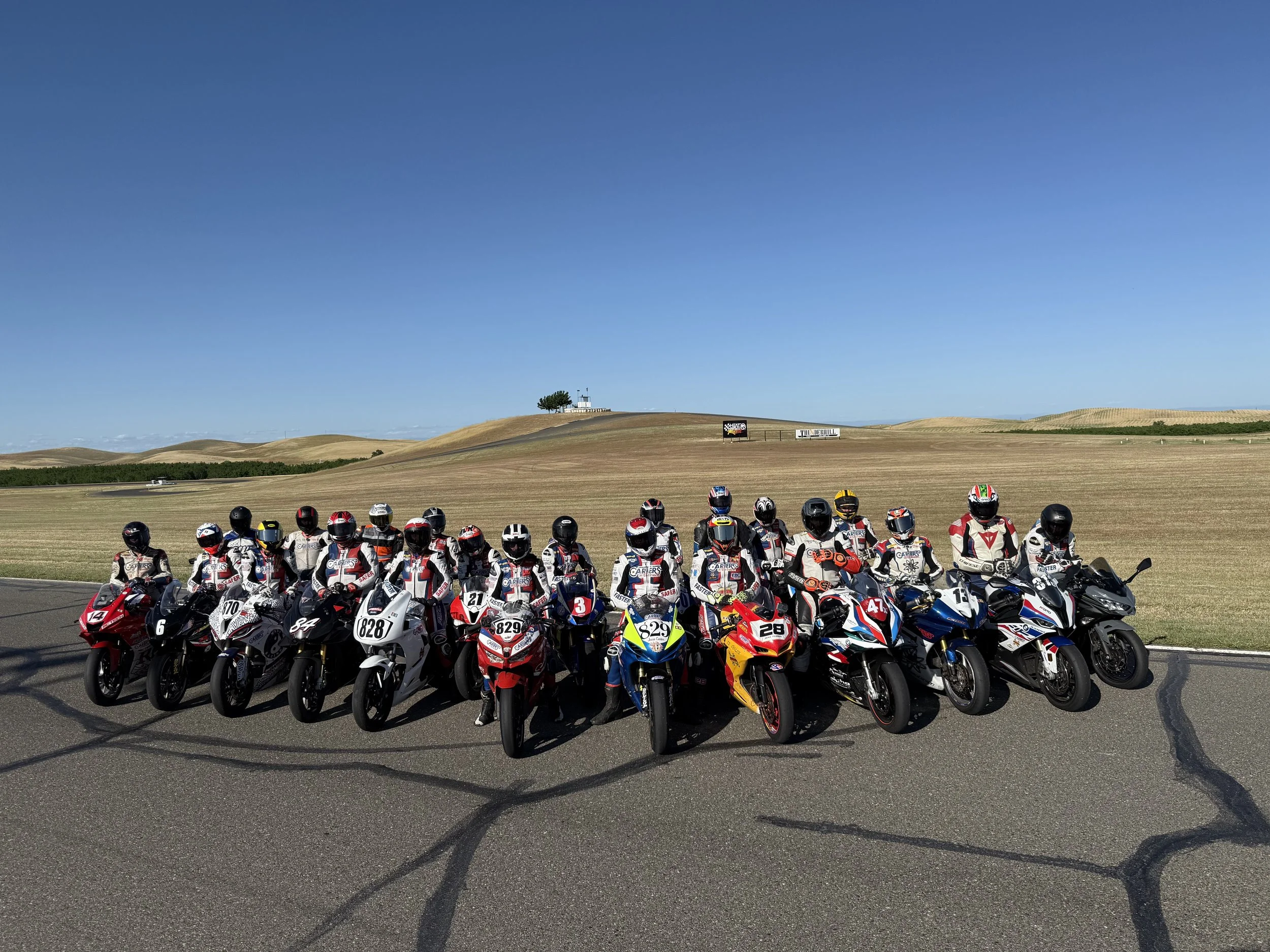 A group of motorcycle racers in racing suits and helmets, lined up on a racetrack, with a highway and open fields in the background under a clear blue sky.