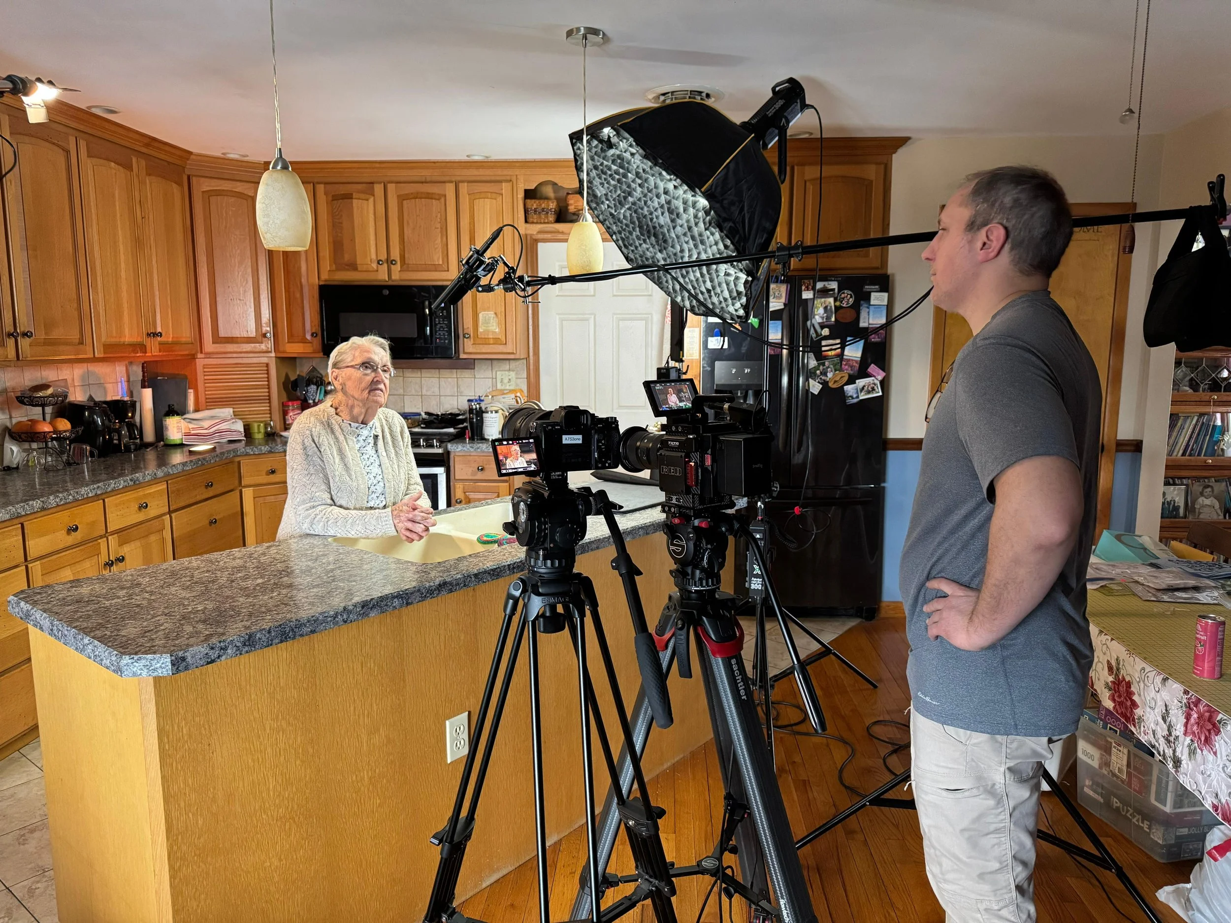 A woman is being filmed by a man in a kitchen. The woman is elderly with white hair, wearing glasses, a light sweater, and sitting at a kitchen island. The man is standing nearby with his hands on his hips, operating video equipment including a camera, microphone, and lighting. The kitchen has wooden cabinets and various appliances.