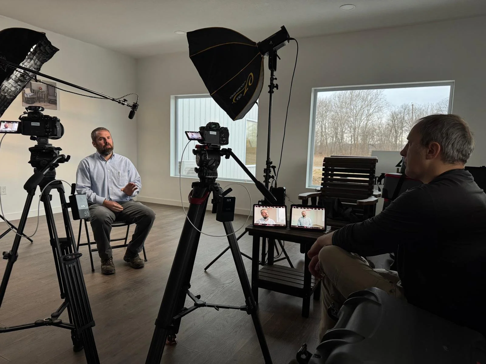 A man in a white shirt sits on a chair in a room, being filmed during an interview or presentation. There are multiple cameras, a lighting setup, and a person with gray hair operating equipment. The room has large windows showing an outdoor scene with trees.