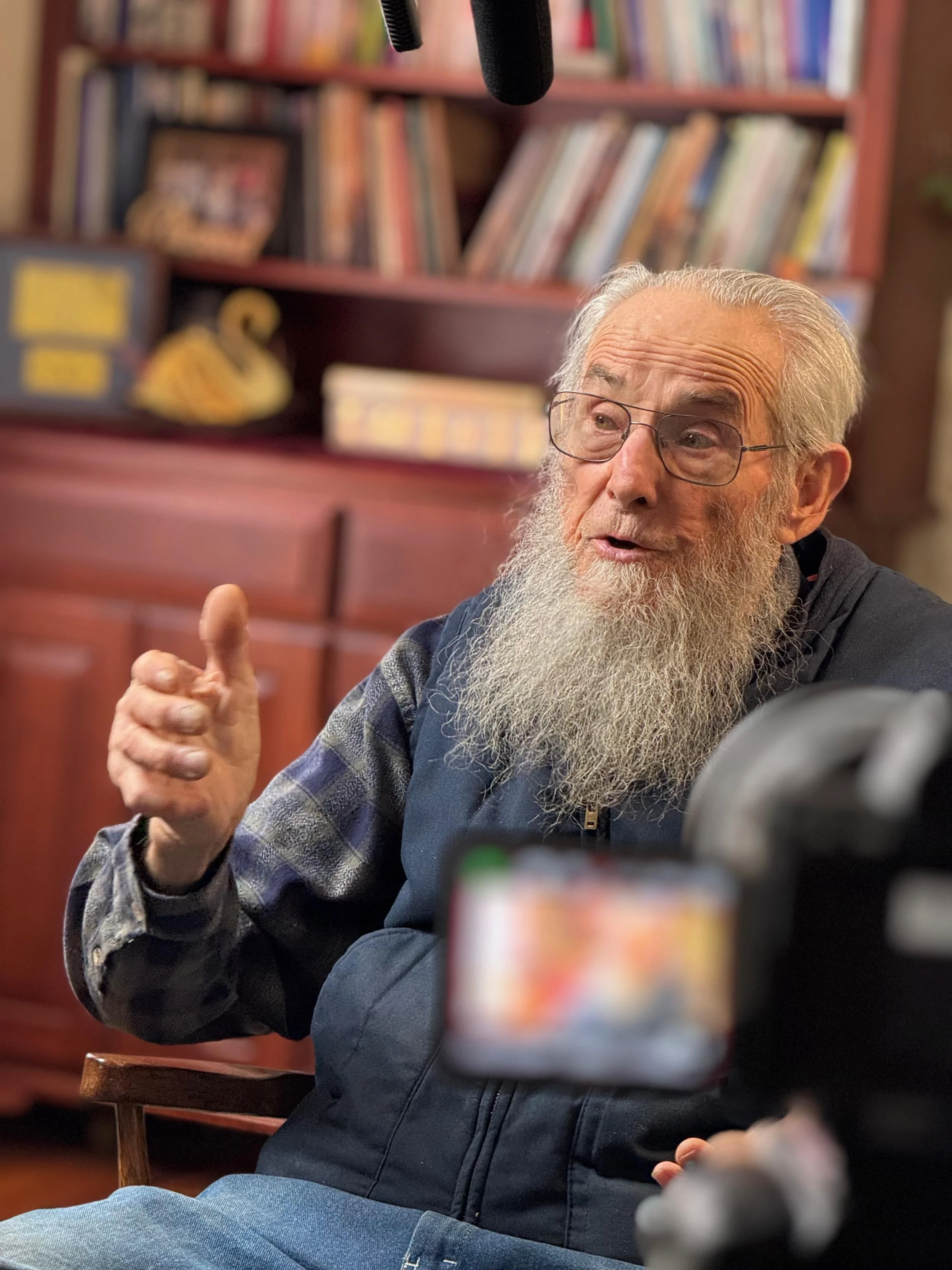 An elderly man with glasses and a long white beard, gesturing with his right hand while speaking, sitting in front of a bookshelf.