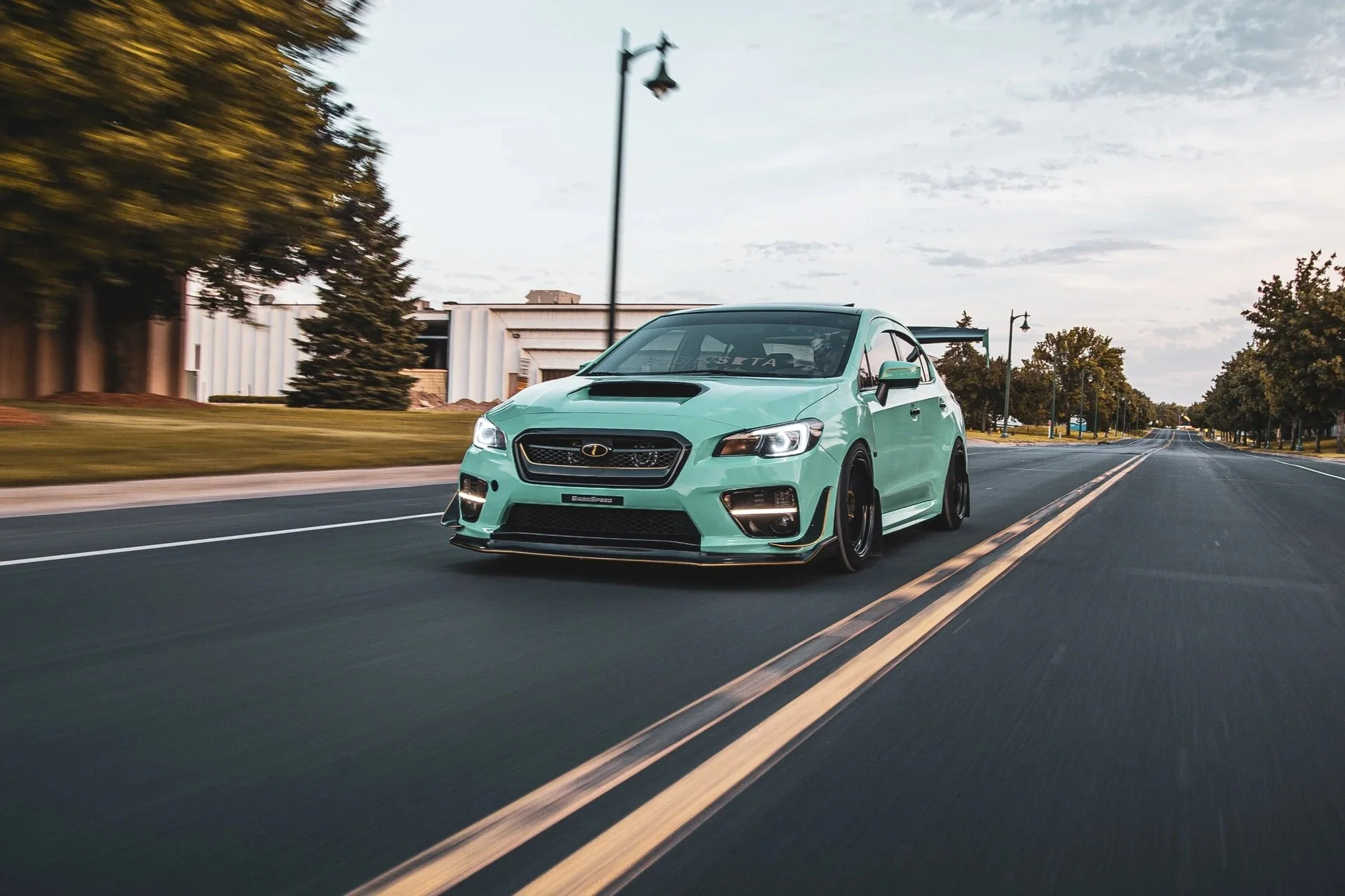 A turquoise Subaru car with a black front grille and black wheels drives on an empty two-lane road lined with trees and lampposts during daytime. The car features a large rear wing, hood scoop, and aftermarket modifications for racing.