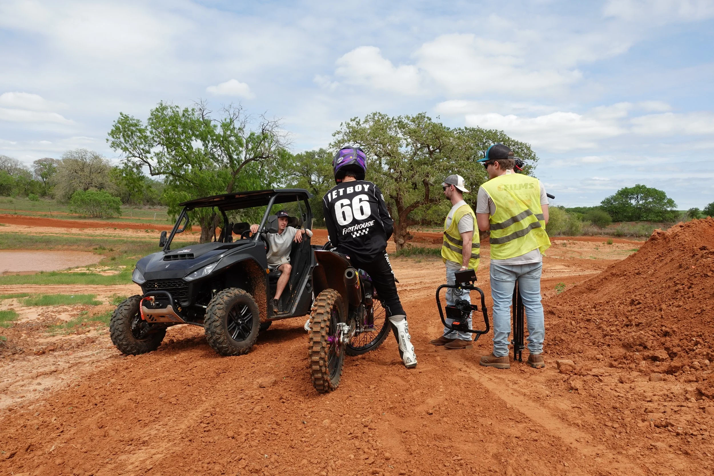 Four men are on a dirt trail surrounded by trees. One man is seated in a side-by-side off-road vehicle, and the others are standing nearby, talking. One of the standing men is wearing a black jersey with the words 'JETBOY 66' on the back, and a purple helmet. Two of the men are wearing yellow safety vests, sunglasses, and hats. The sky is partly cloudy.