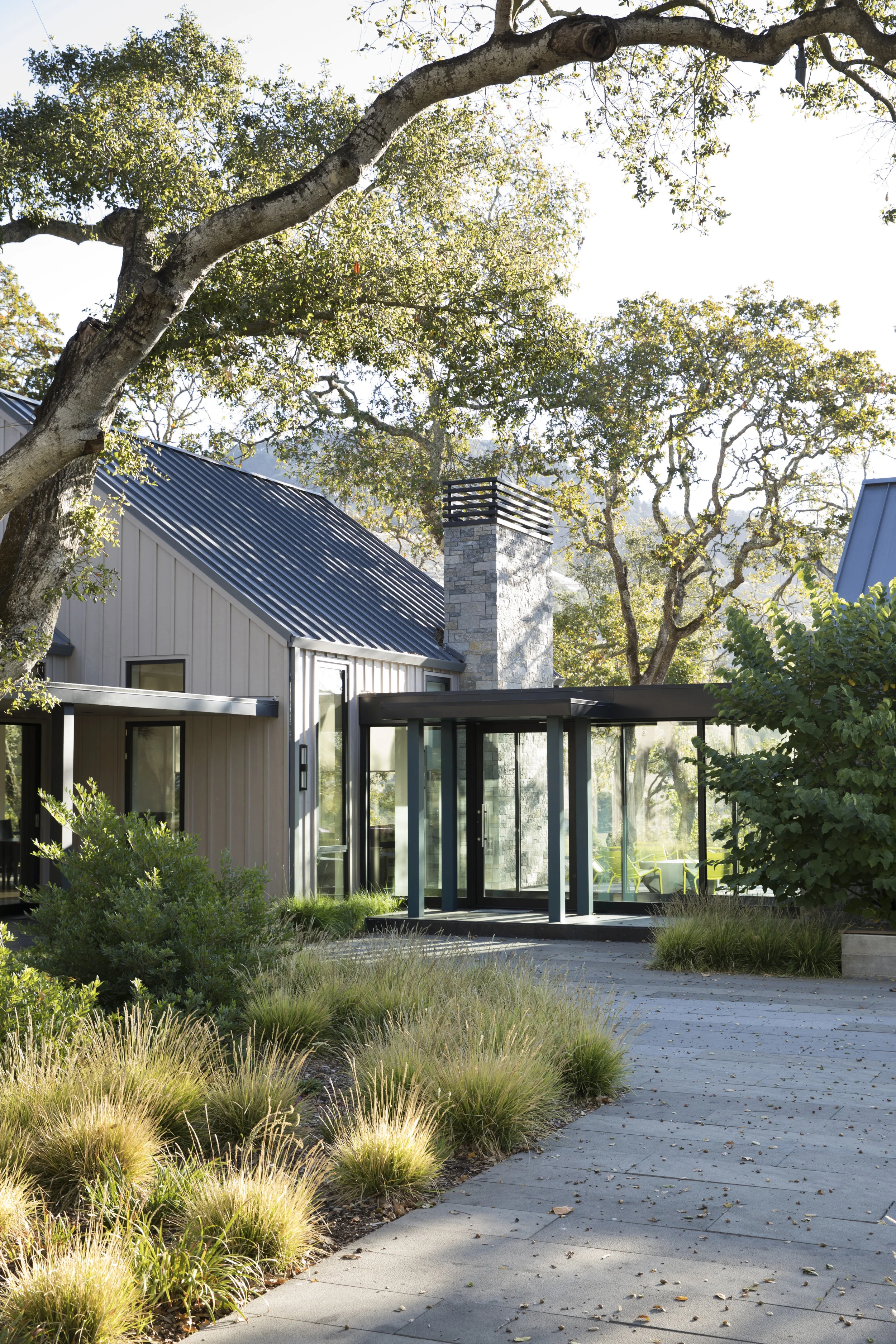 site planting and tile paver hardscape leading to the glass breezeway of a modern sonoma county residence at bennett valley with board and batten siding and a stone veneer chimney
