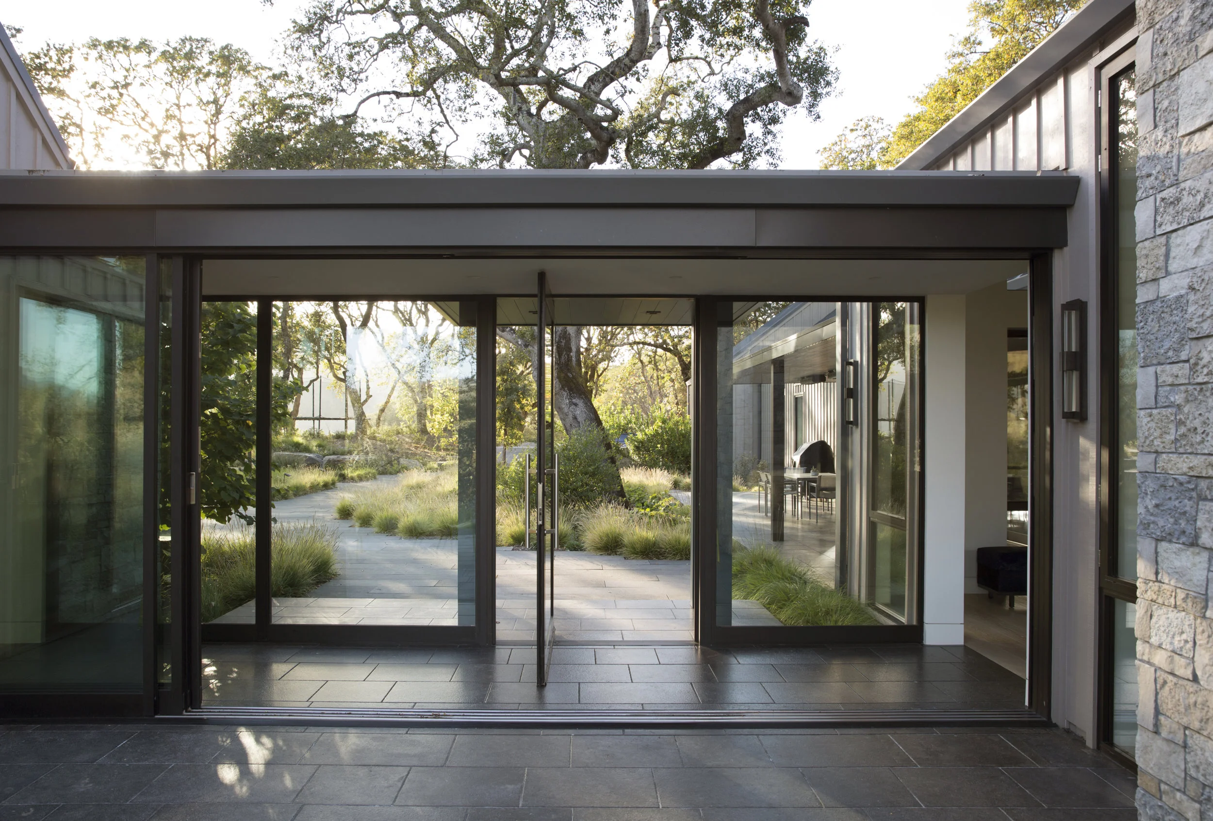 minimalist glass circulation breezeway with large pivot door and sliding glass doors revealing oak trees and tile paving at a modern sonoma county residence in bennett valley