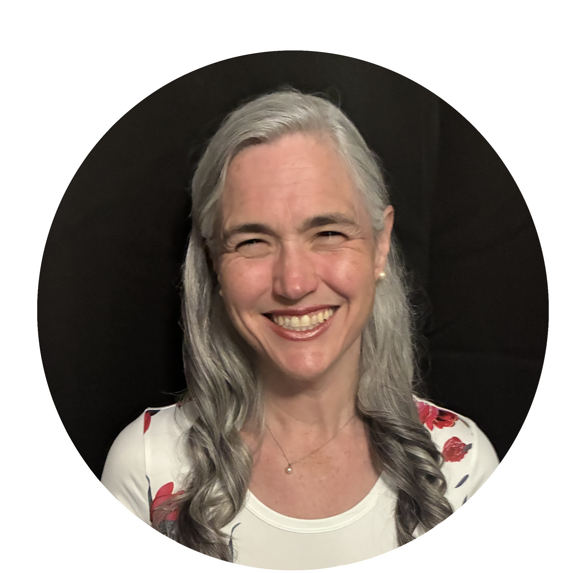 Smiling woman with gray hair, wearing a white top with red and blue floral pattern, and a small necklace, standing in front of a dark background.
