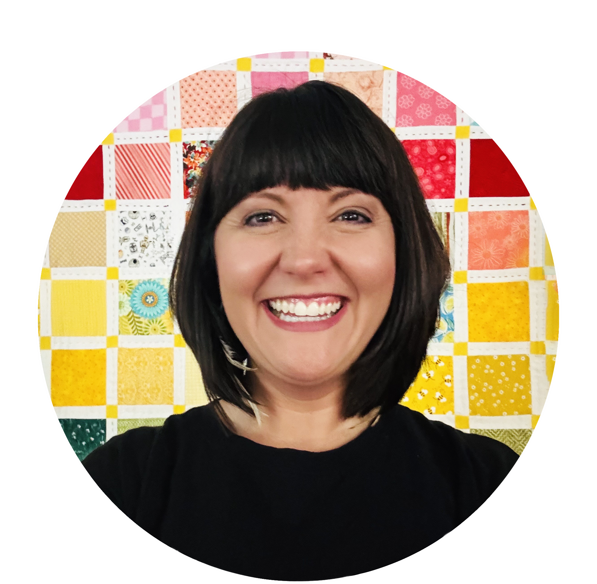 A woman with shoulder-length dark hair and bangs happy, smiling, in front of a colorful quilted wall hanging.