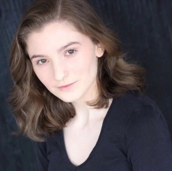 Portrait of a young person with wavy brown hair wearing a black shirt, looking at the camera, against a dark background.