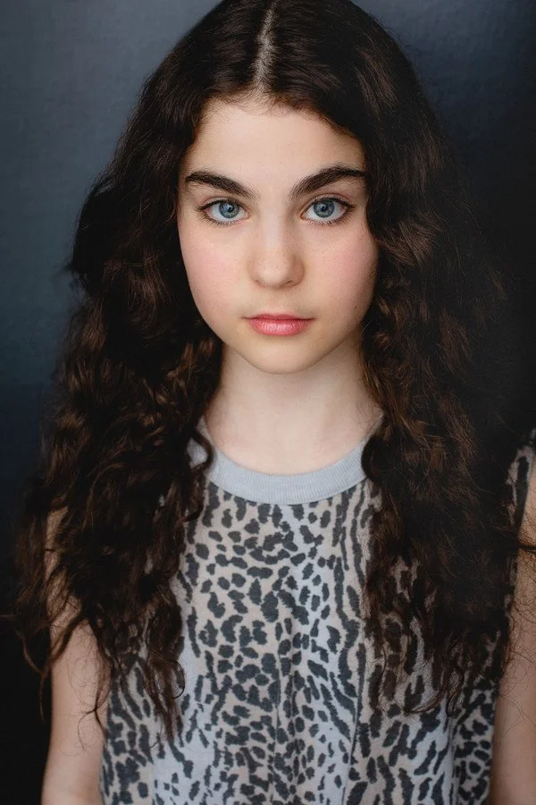 Close-up portrait of a young girl with long, curly dark hair and blue eyes, wearing a leopard print top with a gray collar, against a dark background.