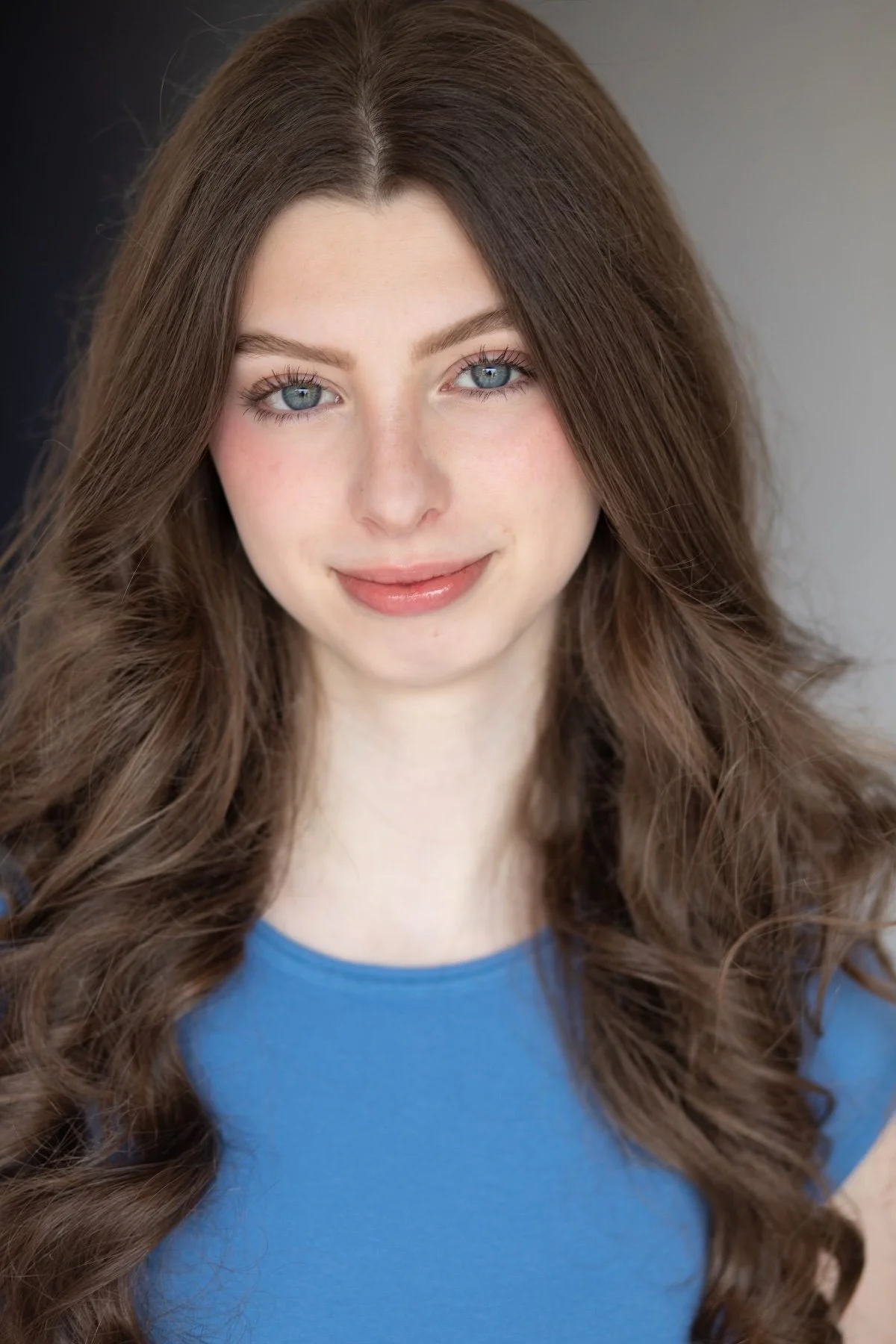 Young woman with long brown hair, blue eyes, and wearing a blue top smiling slightly at the camera.