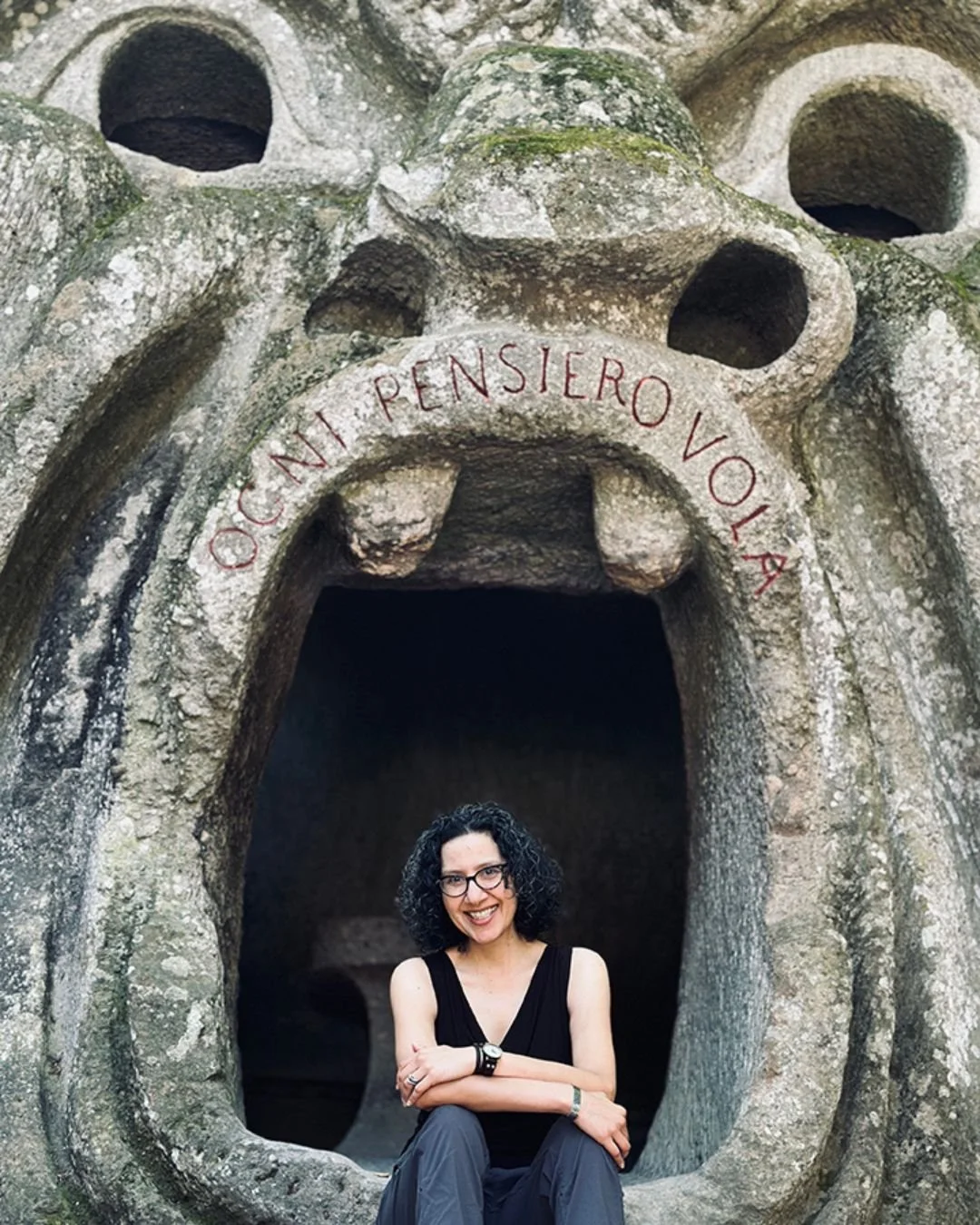 Gigi Pandian sitting in the mouth of a huge stone ogre in Bomarzo, Italy's Sacro Bosco Sacred Wood Park of Monsters.