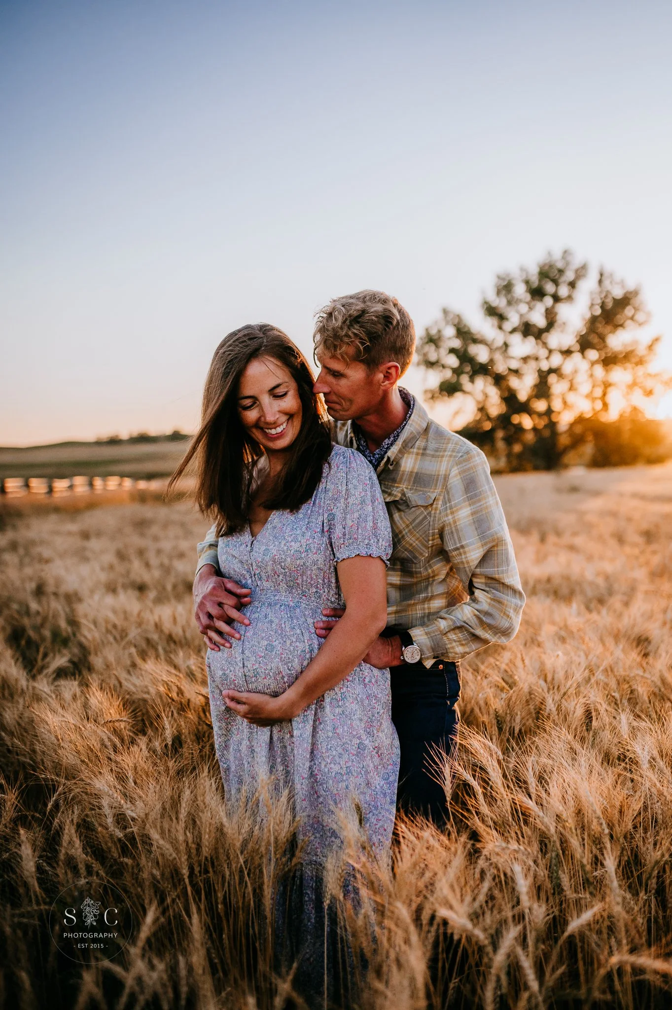 Pregnant woman embraced by her husband in a golden wheat field at sunset, maternity photographer in Belfield, North Dakota