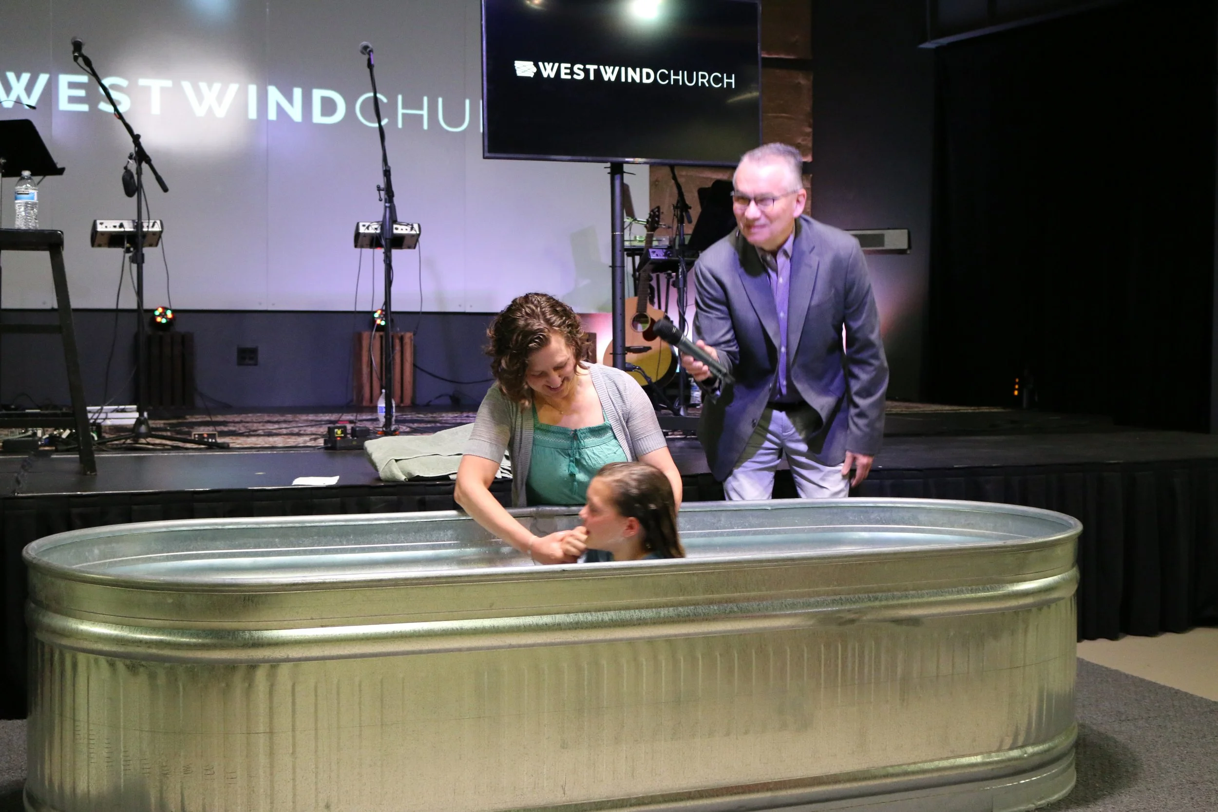 A woman and a young girl are inside a metal baptismal pool at Westwind Church. Behind them, a man in a suit is speaking into a microphone, smiling. The stage has musical instruments, microphones, and a large screen displaying the church's name.