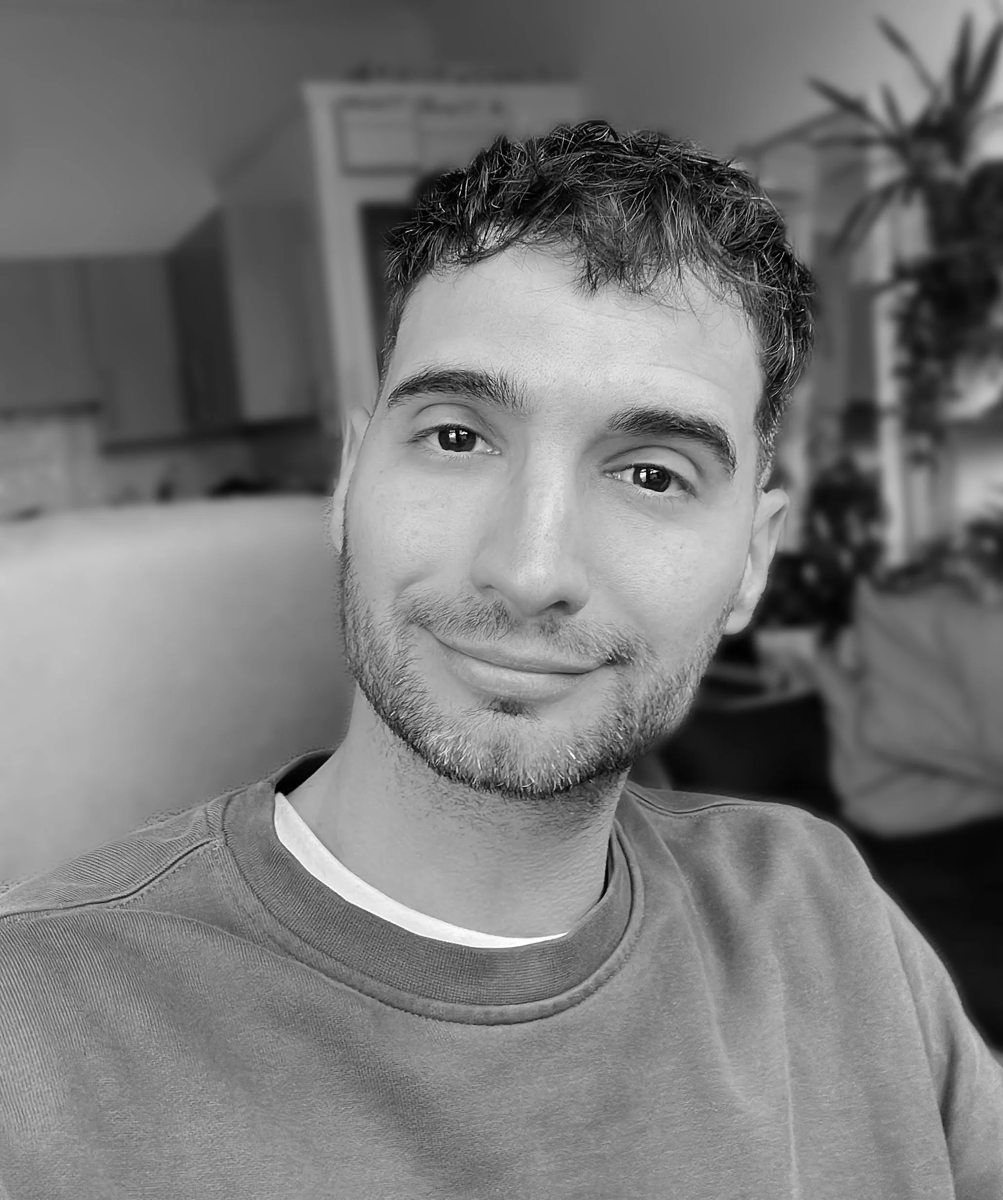 A man with short dark hair and a light beard smiling, sitting indoors in front of blurred background with house plants and furniture, in black and white.