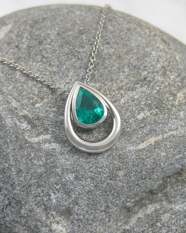 A drop pendant features a vibrant pear shaped emerald set within a smooth rubover setting mounted with a pear shaped halo of platinum.jpg