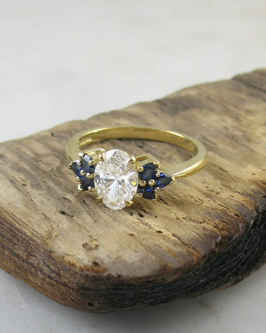 Bespoke oval diamond engagement ring with blue sapphire cluster shoulders in yellow gold