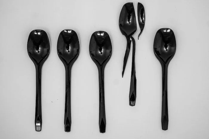 A small collection of 5 black plastic shiny spoons lie against a white plain background, one is broken sitting higher than the rest