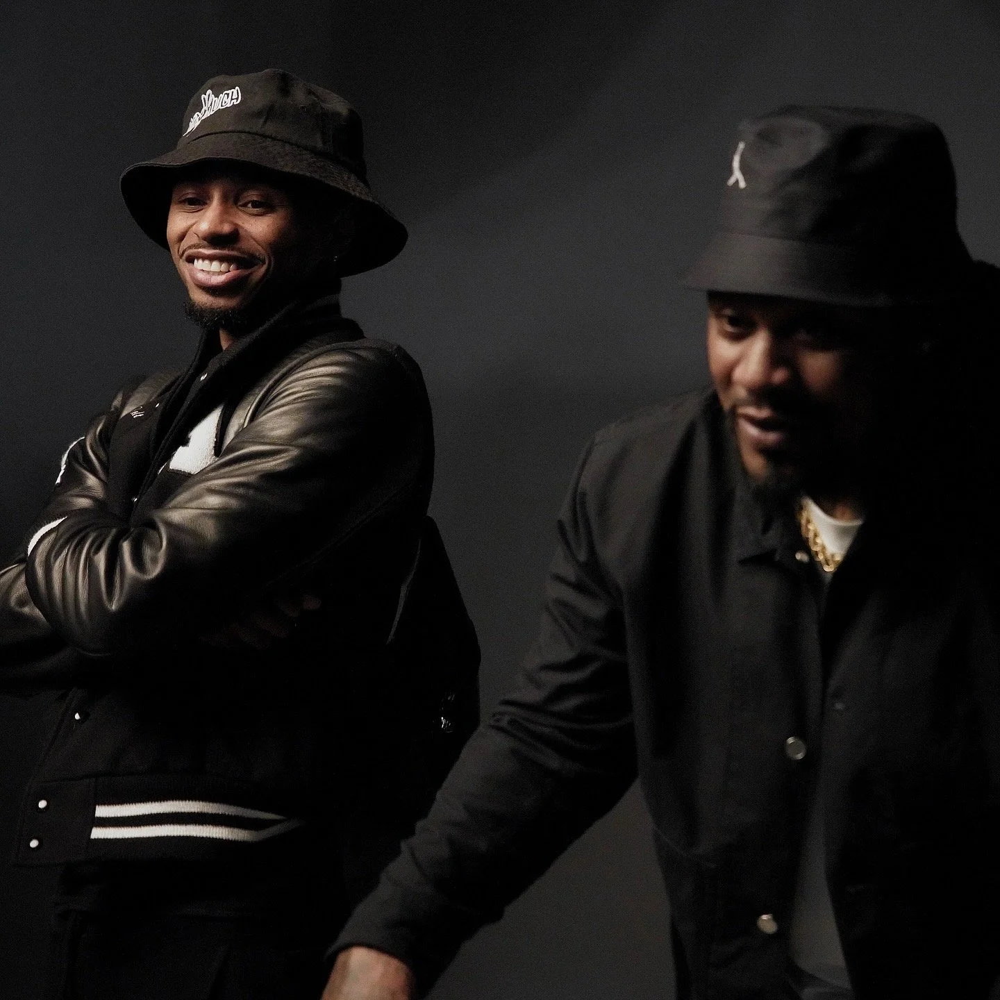 Two men in dark clothing and bucket hats smiling against a black background.