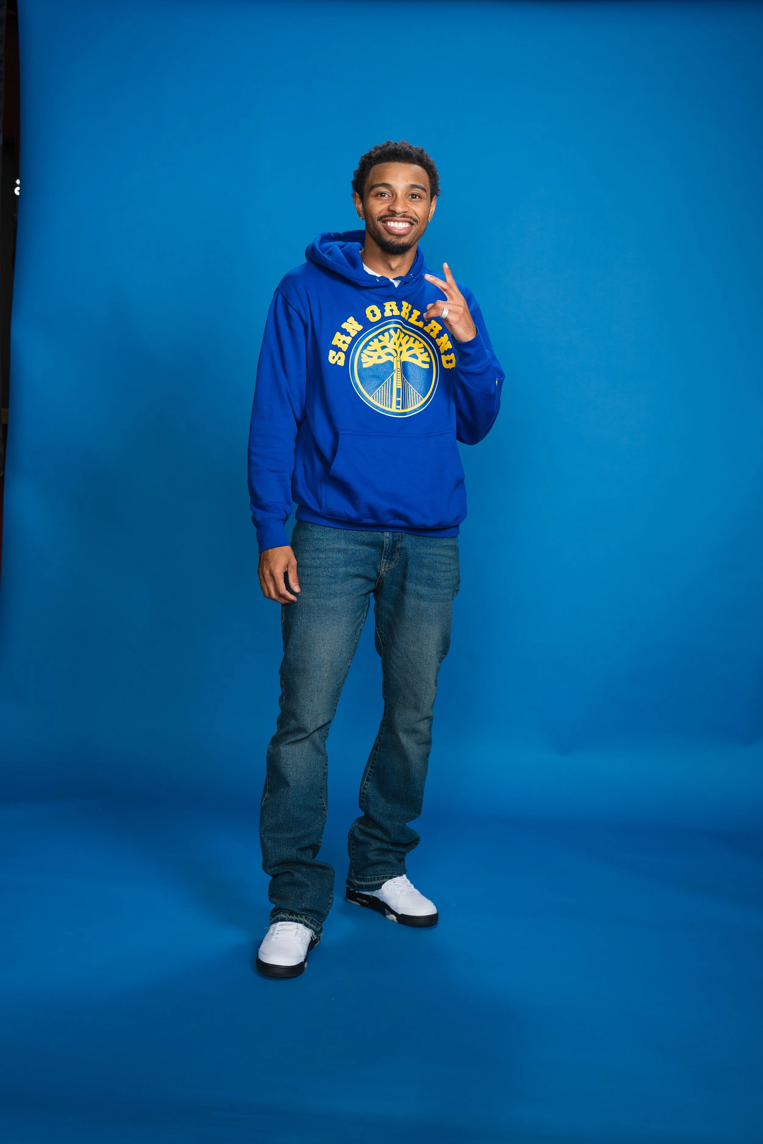 A young man smiling and making a peace sign with his right hand, wearing a blue San Francisco sweatshirt, jeans, and white sneakers, standing against a solid blue background.