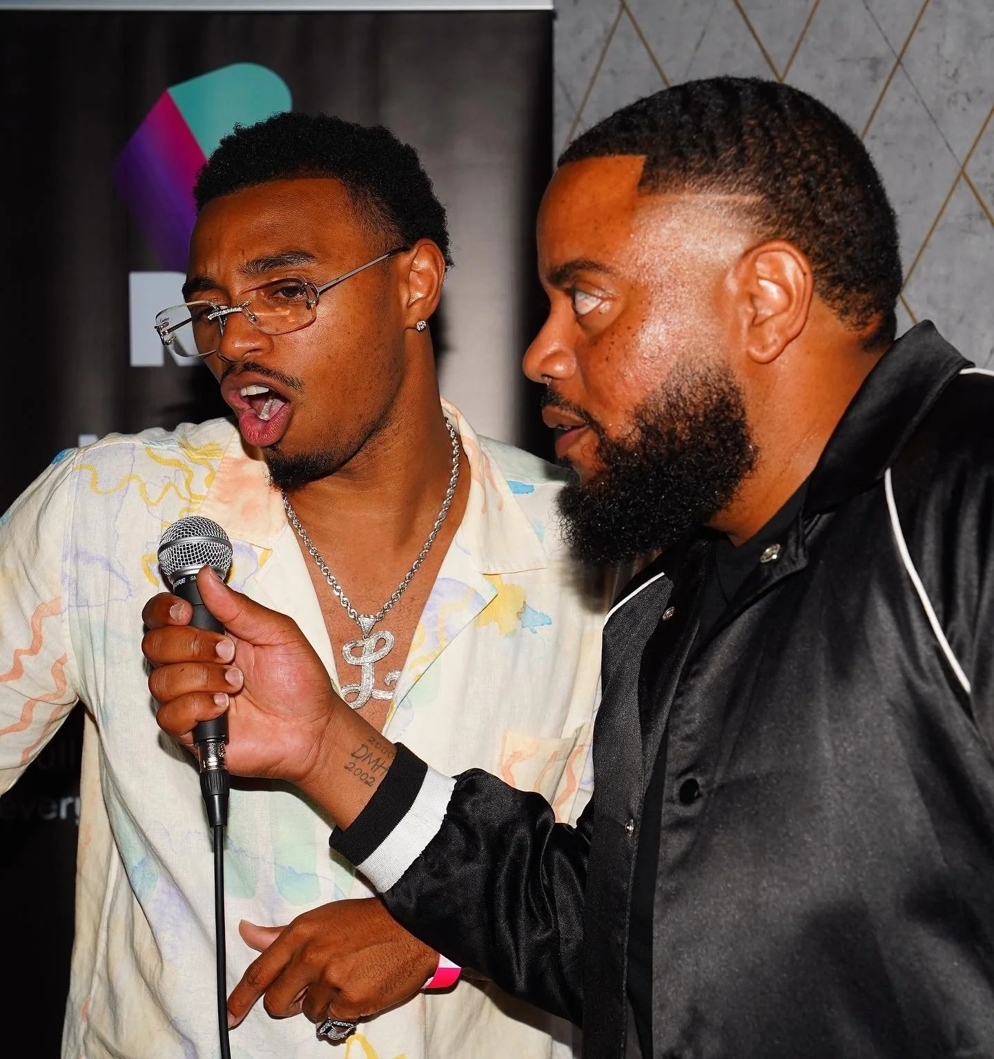 Two men are engaged in a conversation, one holding a microphone. The man on the left wears glasses and a colorful shirt with a necklace, while the man on the right has a beard and wears a black jacket.