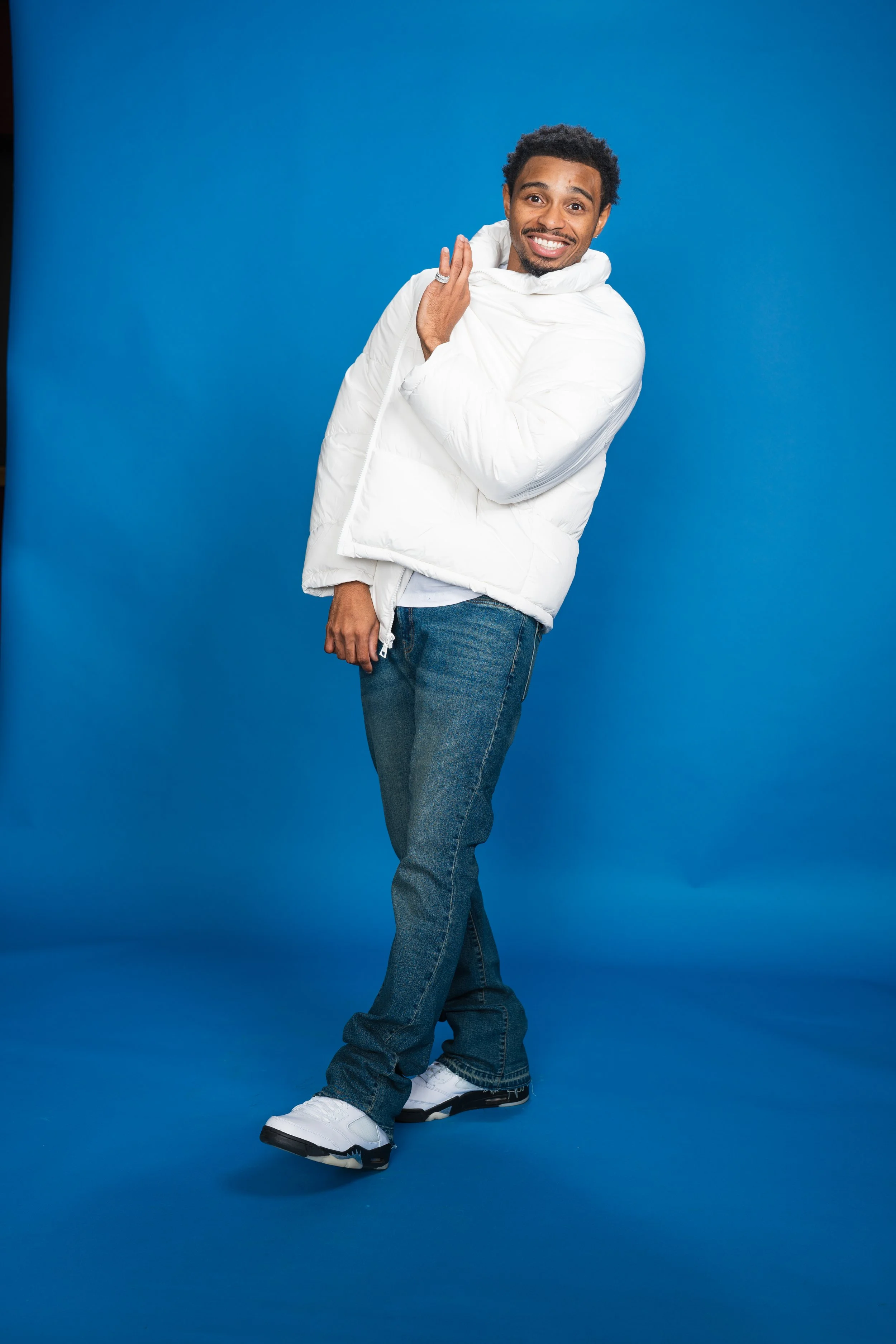 A smiling man wearing a white puffer jacket, jeans, and white sneakers standing against a blue background.