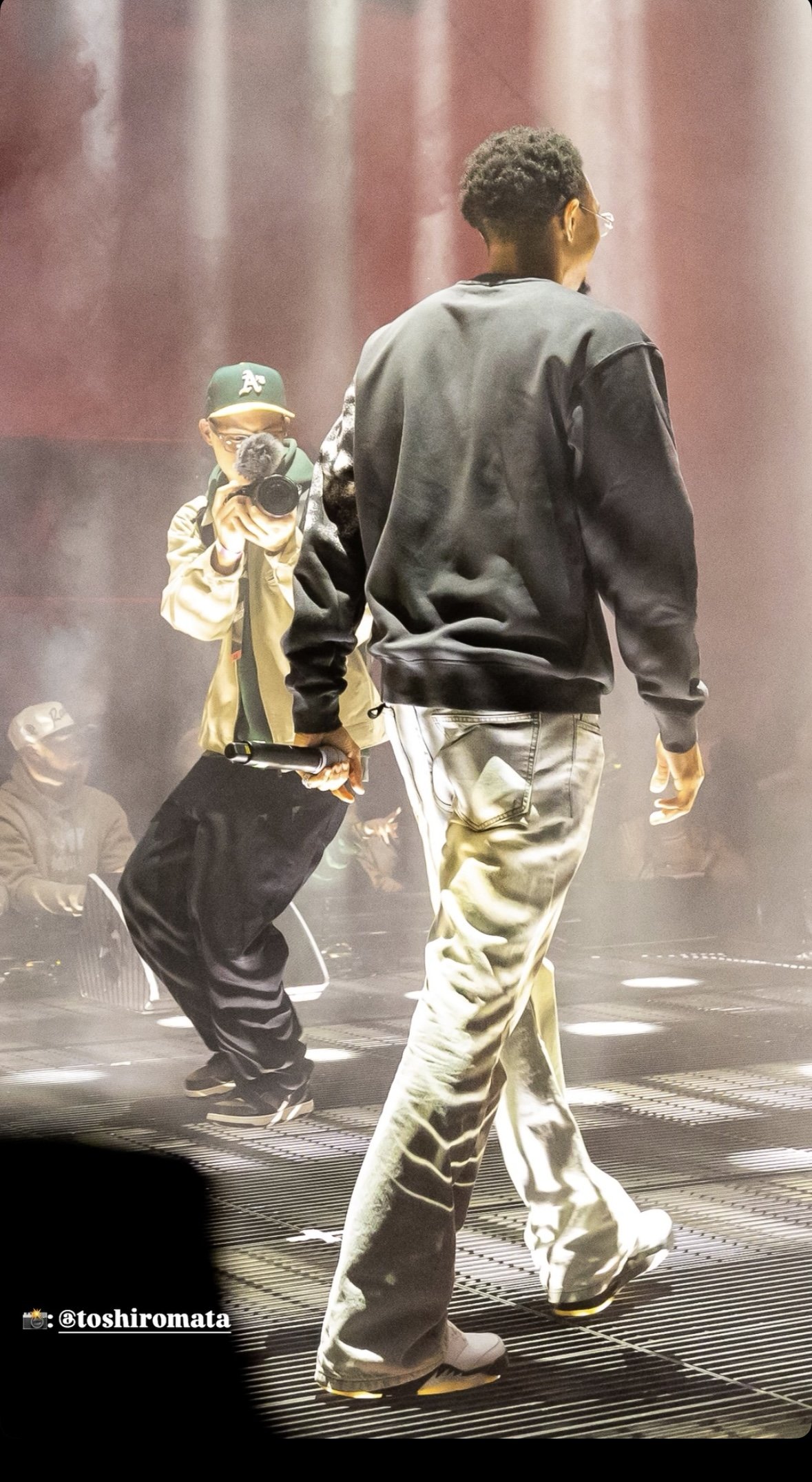 A person walking on stage with a performer behind is holding a camera and microphone, wearing a tan jacket and black cap, stage fog and lighting in the background.