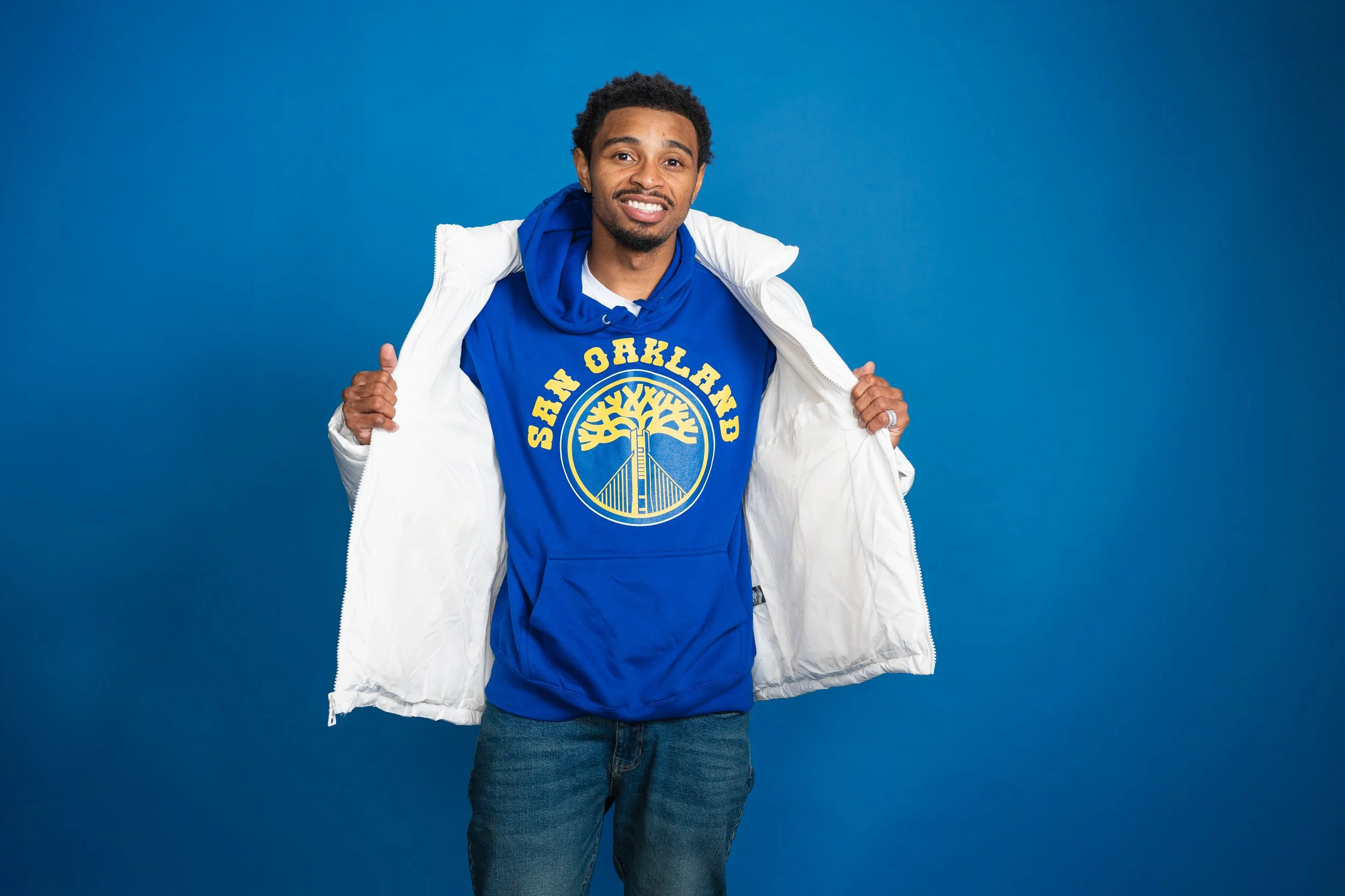 A man wearing a blue hoodie with a yellow emblem and text, holding a white jacket open to reveal the hoodie, standing against a solid blue background.